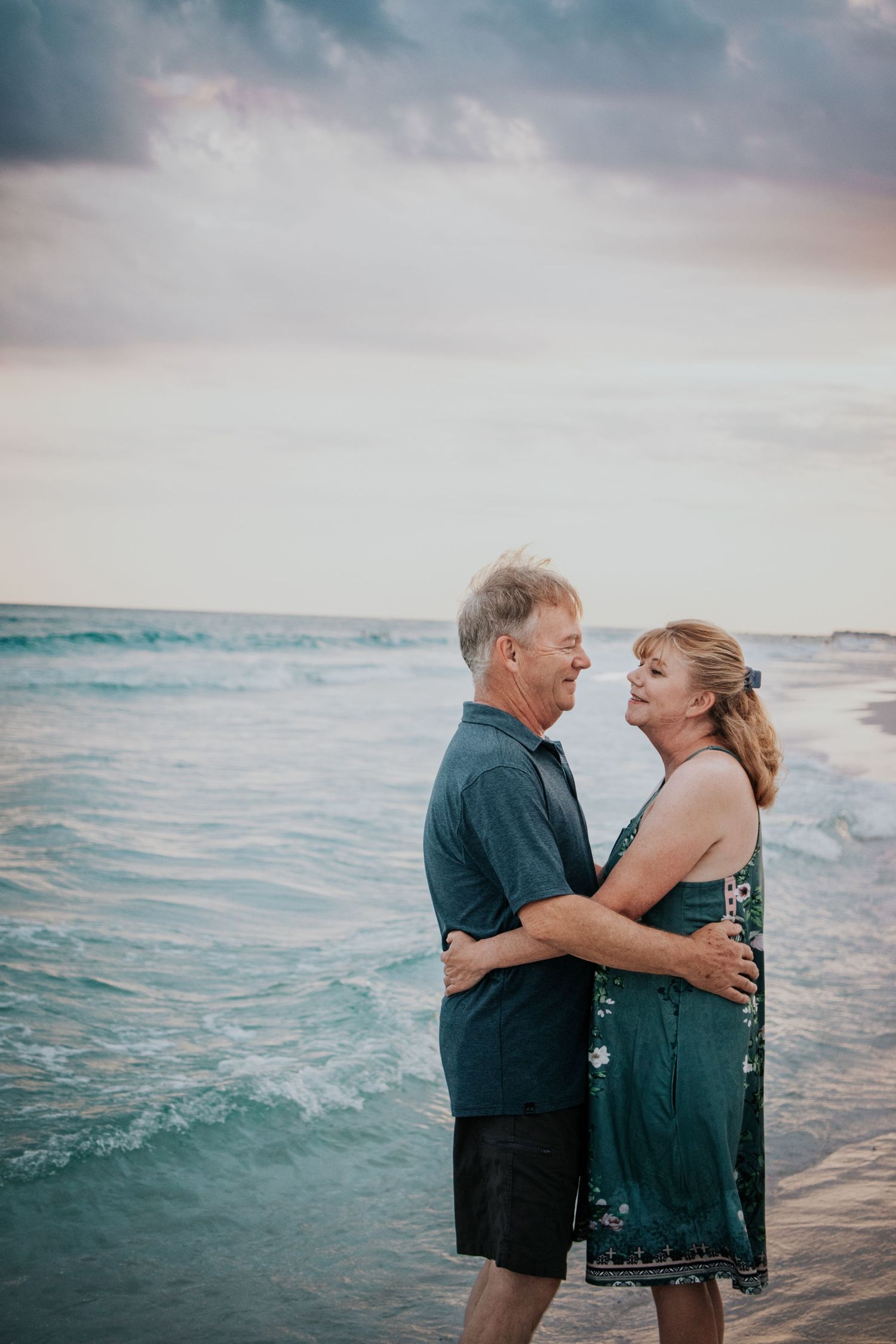 Middle-aged couple holding each other close while standing in the ocean surf at sunset.