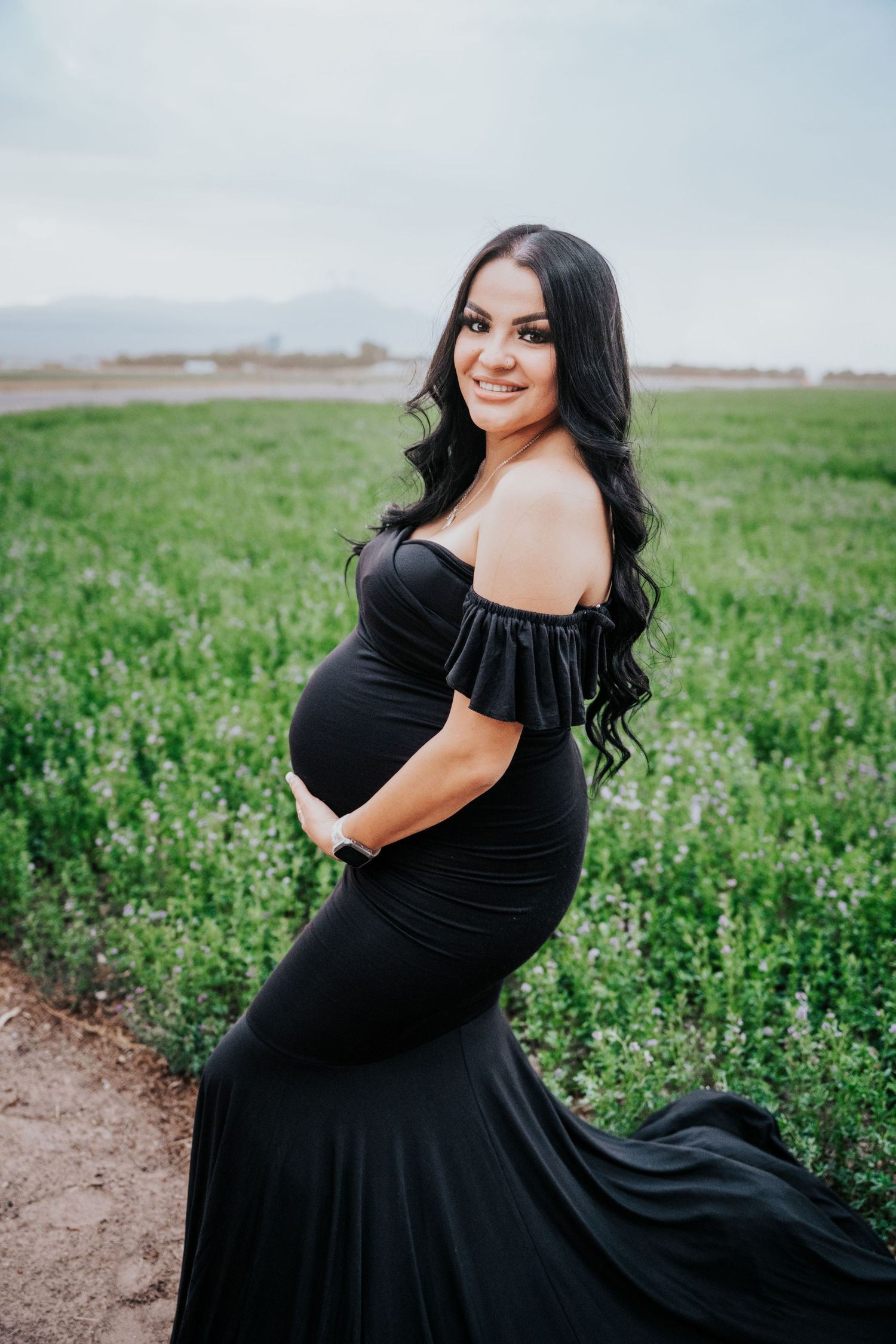 Radiant pregnant woman in black maternity gown with flowing train poses in green field with mountains.