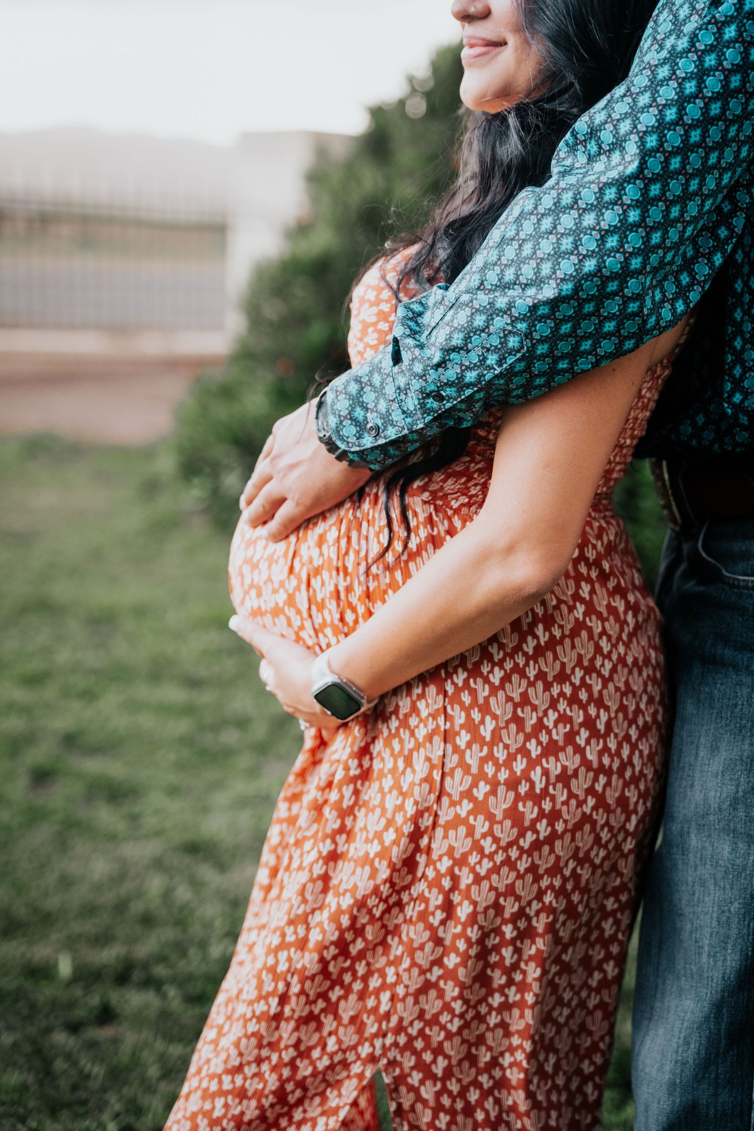 Man in teal shirt embracing pregnant woman in orange floral dress from behind, highlighting her baby bump.