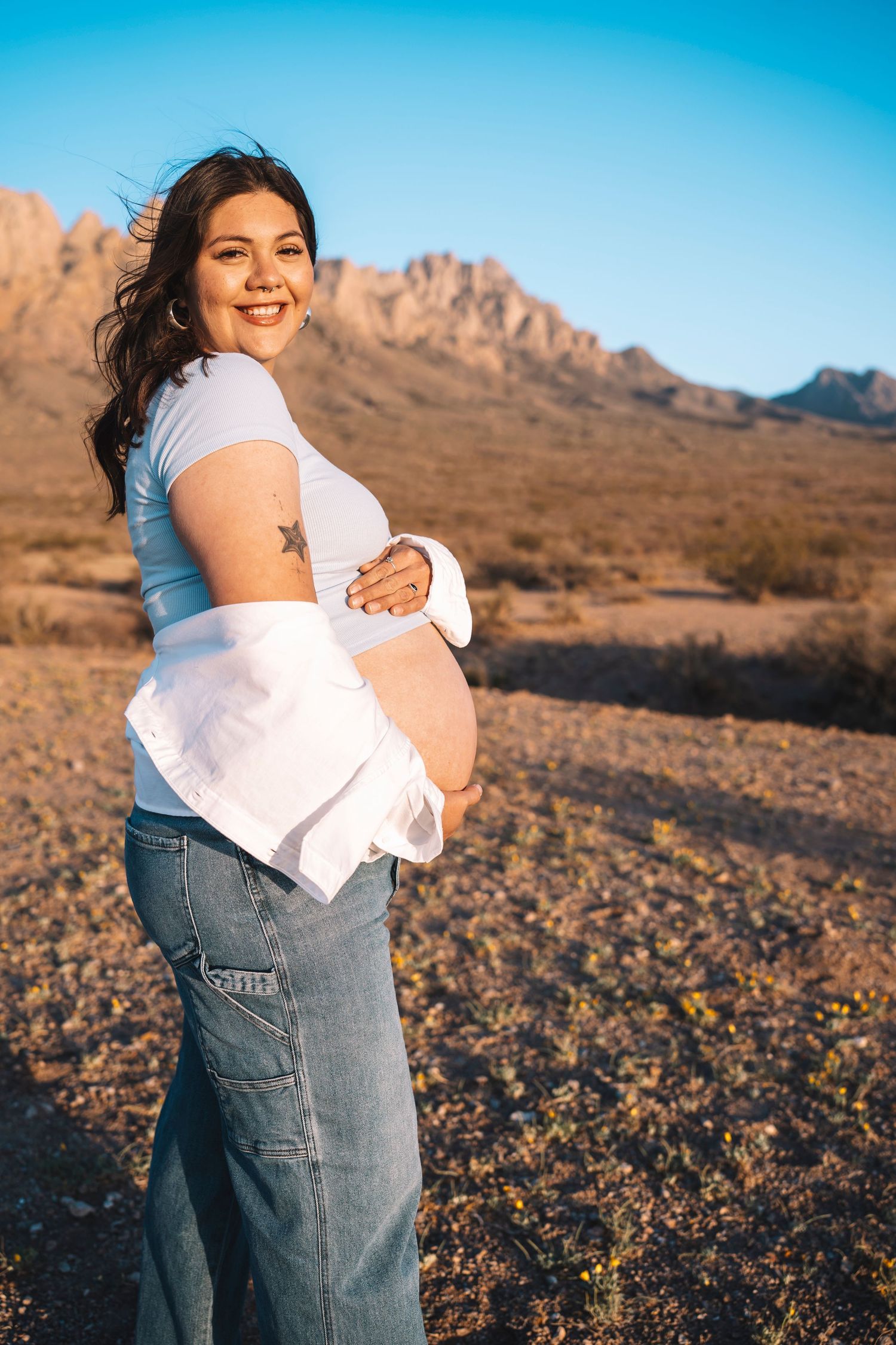 Smiling pregnant woman in white top and jeans cradling baby bump outdoors with desert mountains at golden hour.