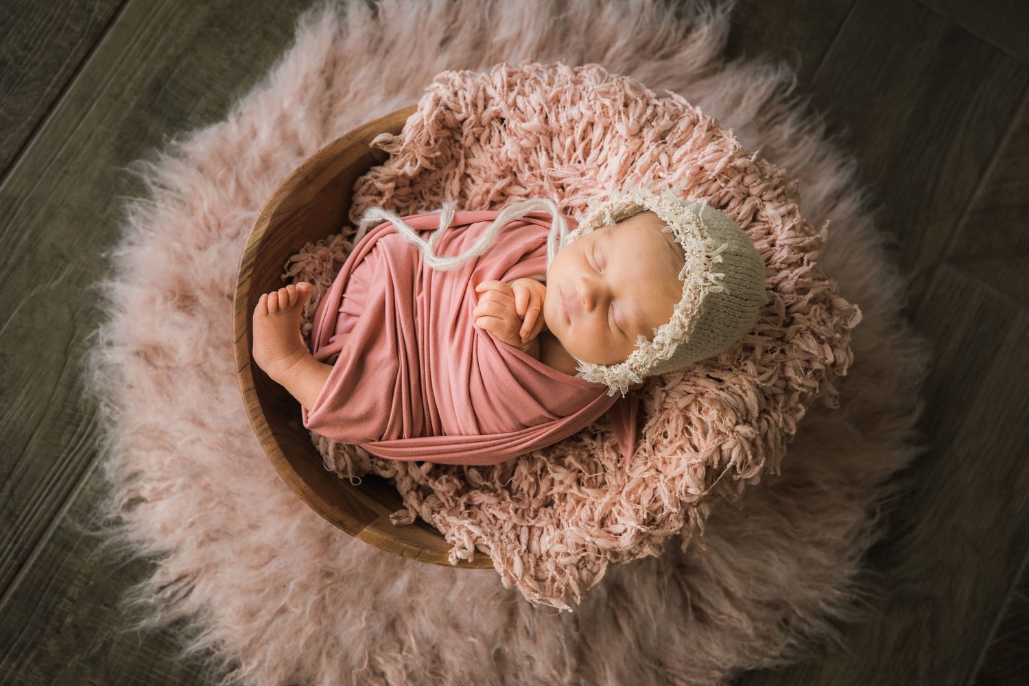 Overhead view of newborn baby wrapped in pink swaddle wearing lace bonnet in a wooden bowl.