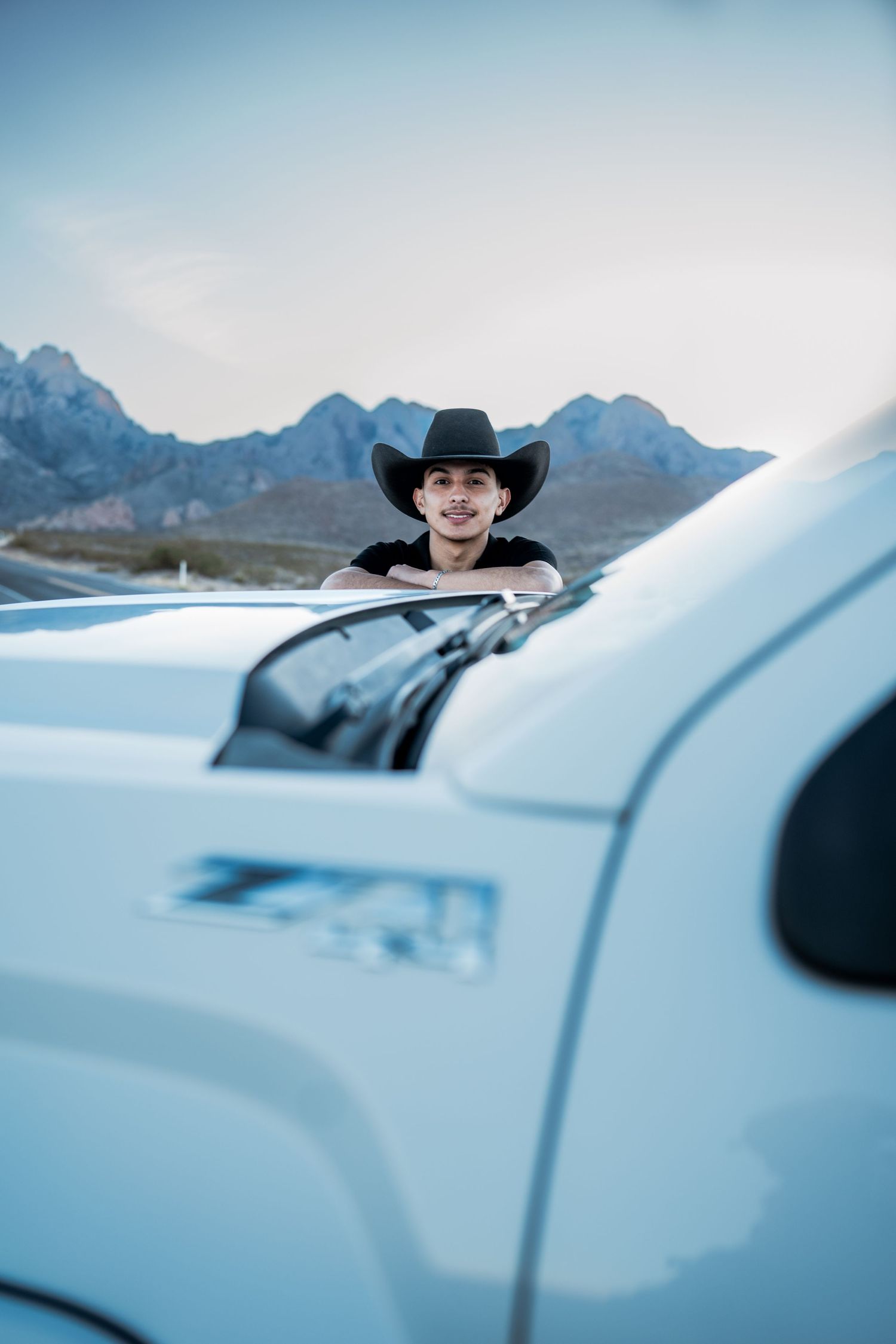 Smiling young man in black cowboy hat leaning over white truck with mountain backdrop.