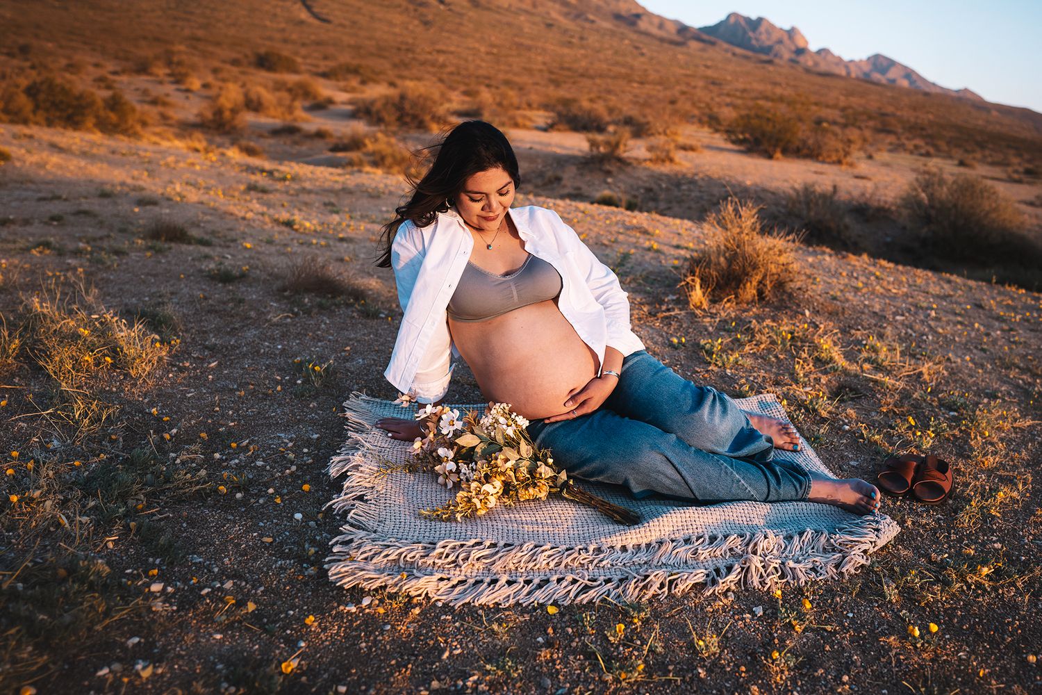 Happy pregnant woman sitting on blanket holding wildflowers and baby bump in desert sunset.