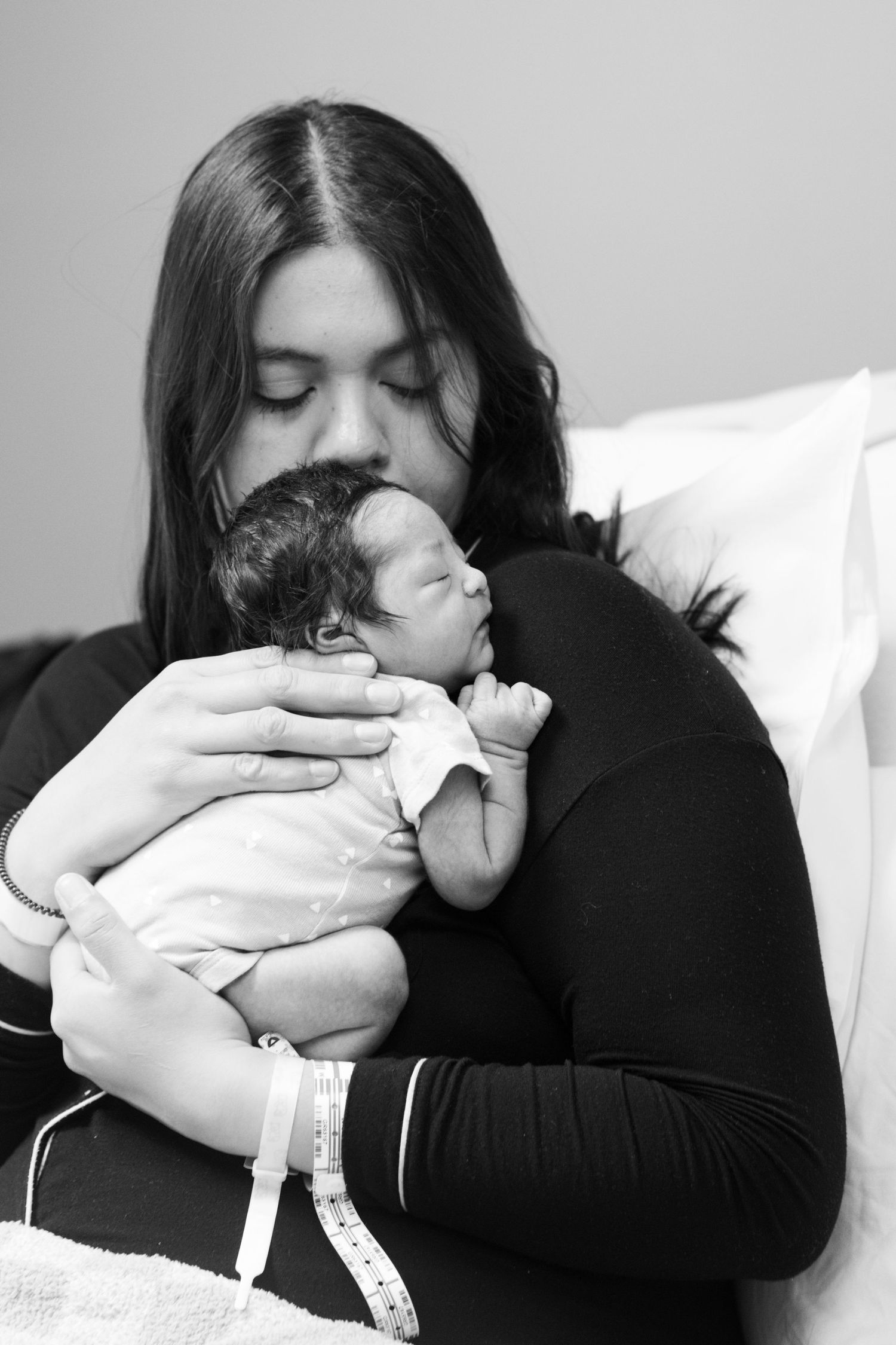 Black and white portrait of new mother with hospital bracelet kissing her sleeping newborn baby.