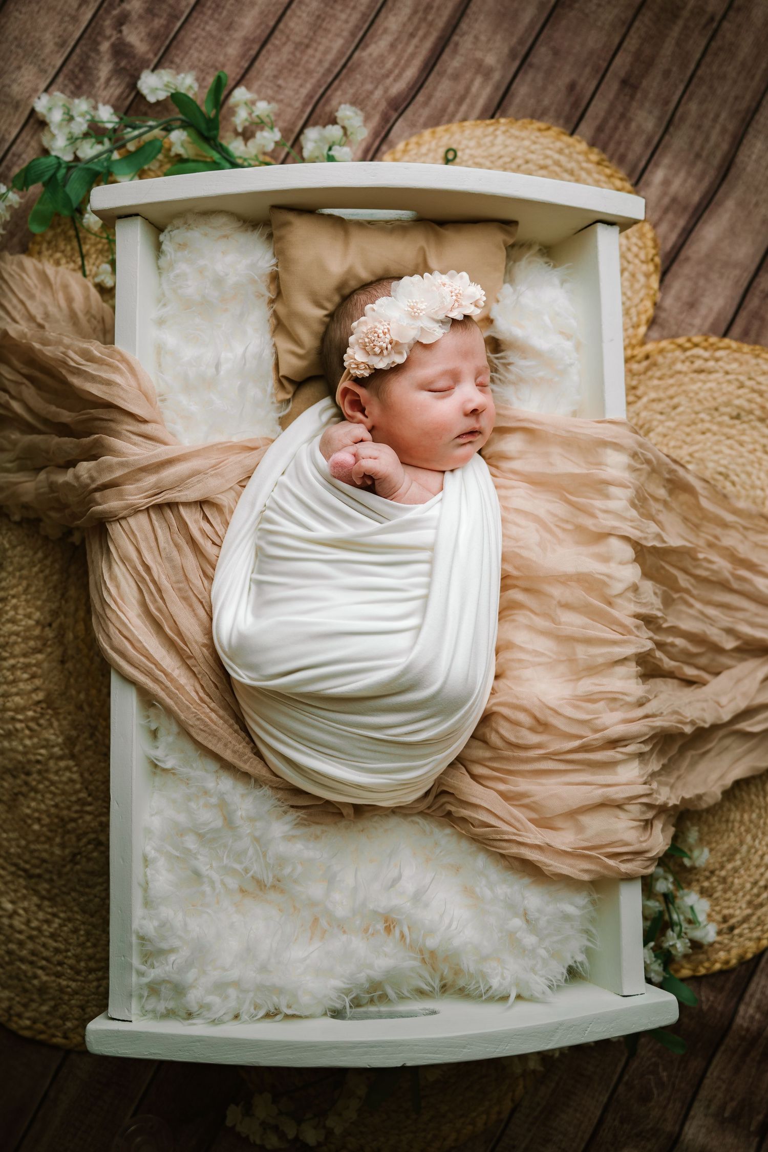Sleeping newborn baby girl wrapped in white swaddle with floral headband in a small white wooden bed.
