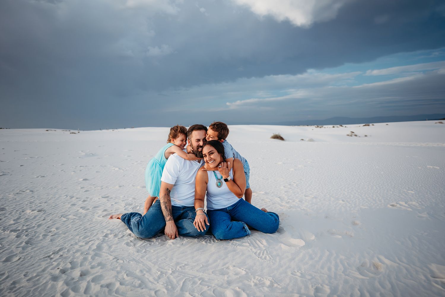 Loving family of four cuddling closely together while sitting on white sand dunes outdoors.