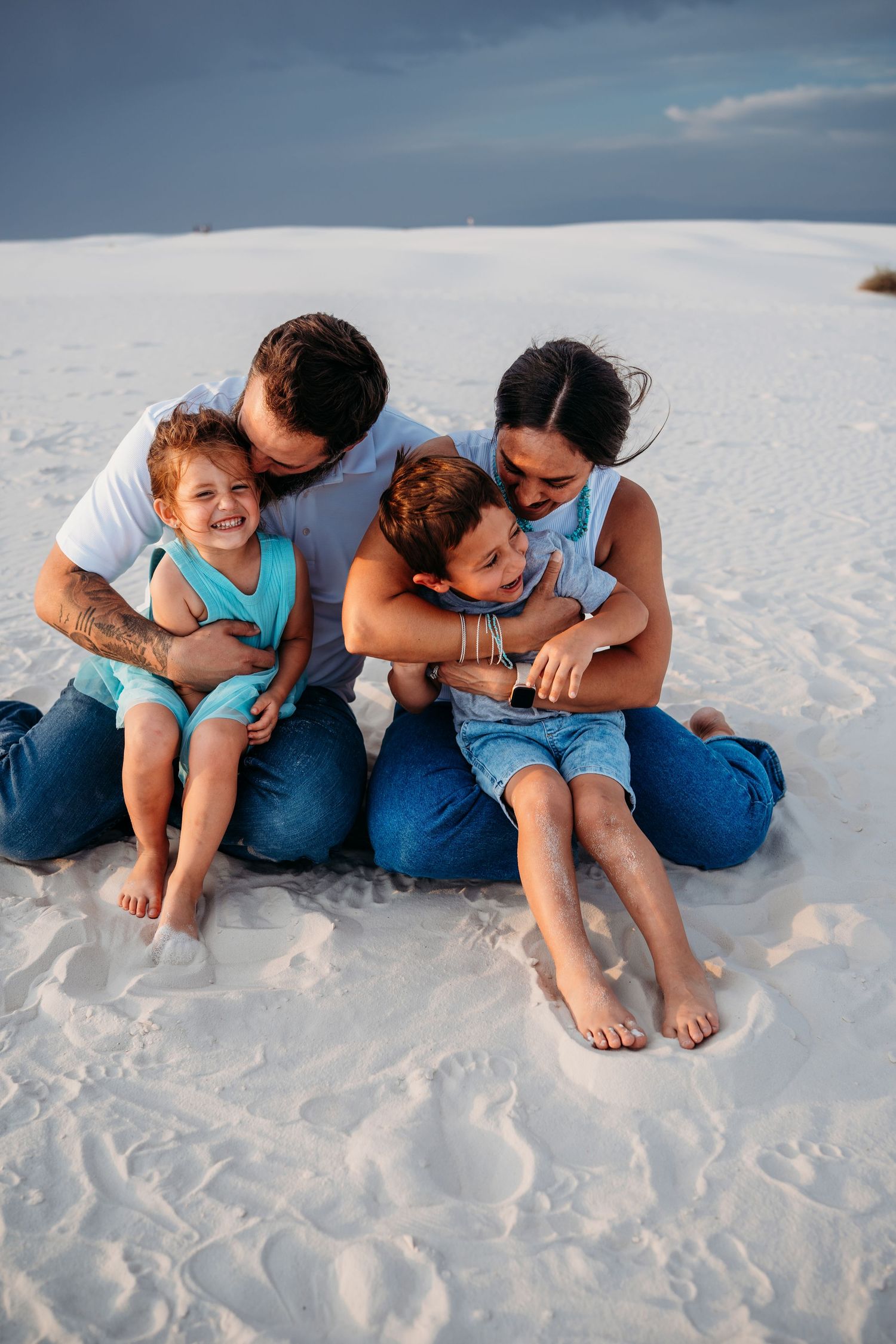 A loving family of four cuddles and laughs together while sitting on bright white sand dunes.