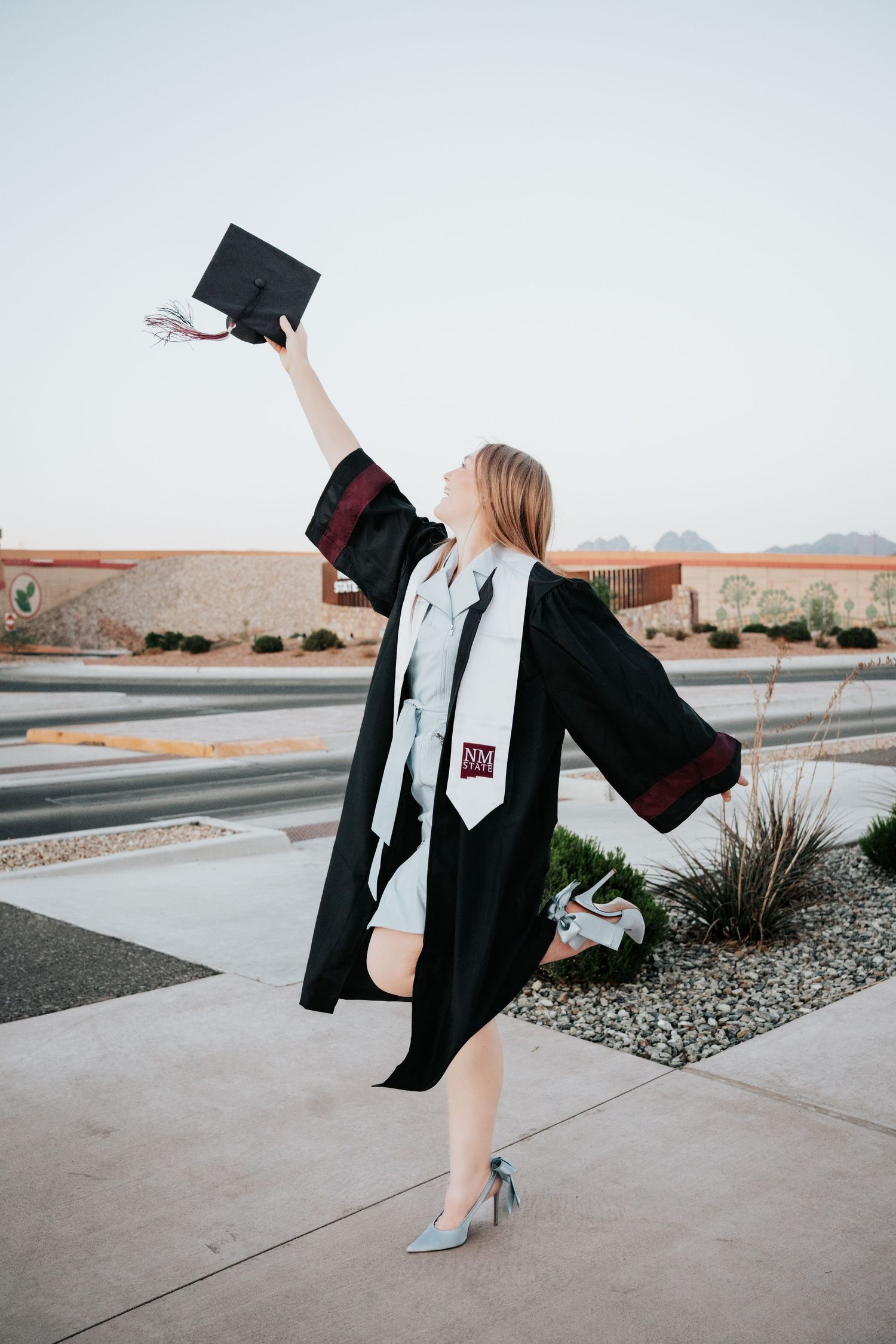 A female graduate in black cap and gown raises her mortarboard triumphantly outdoors at New Mexico State University.