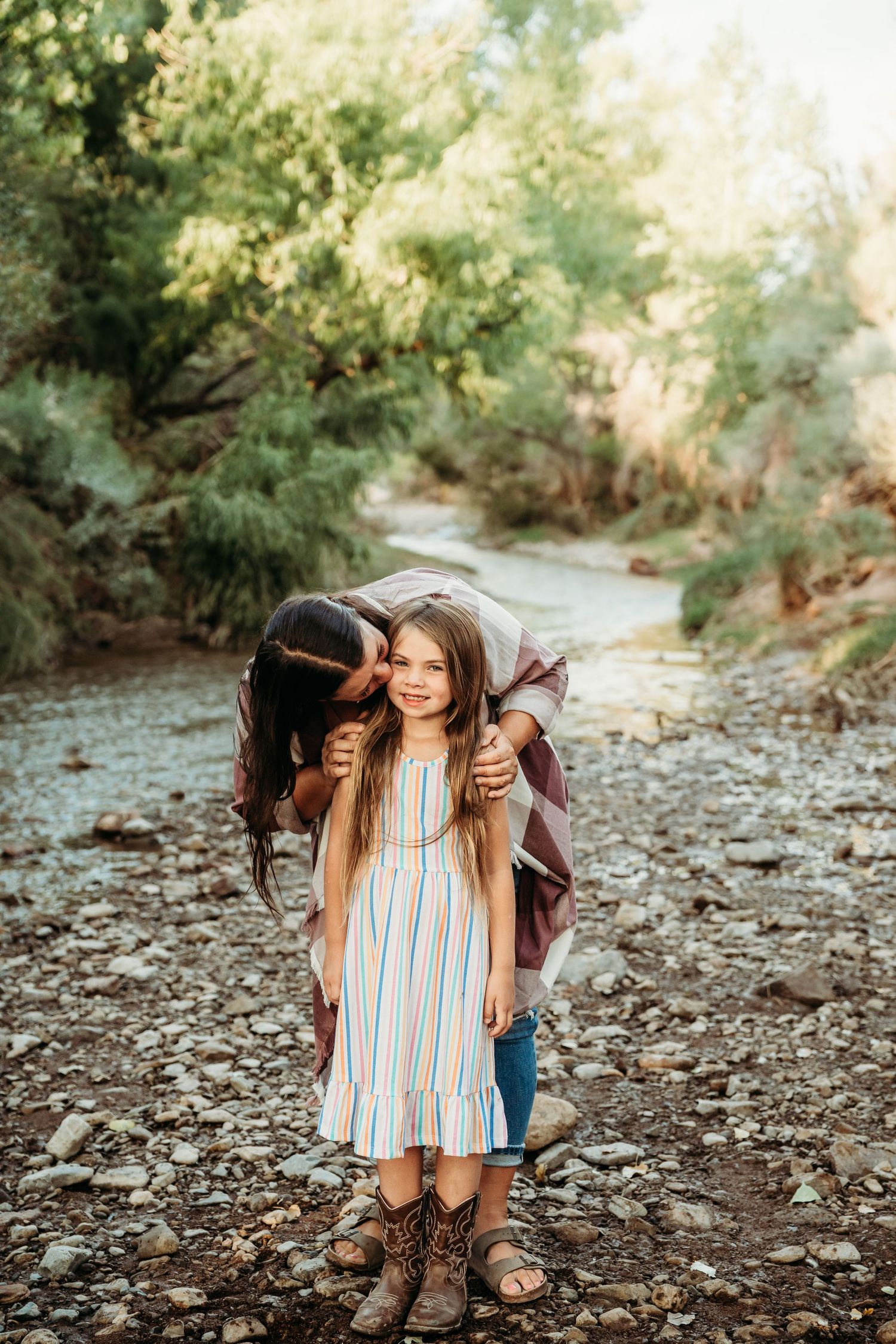 A woman kisses a smiling young girl in a striped dress outdoors near a rocky creek surrounded by green trees.