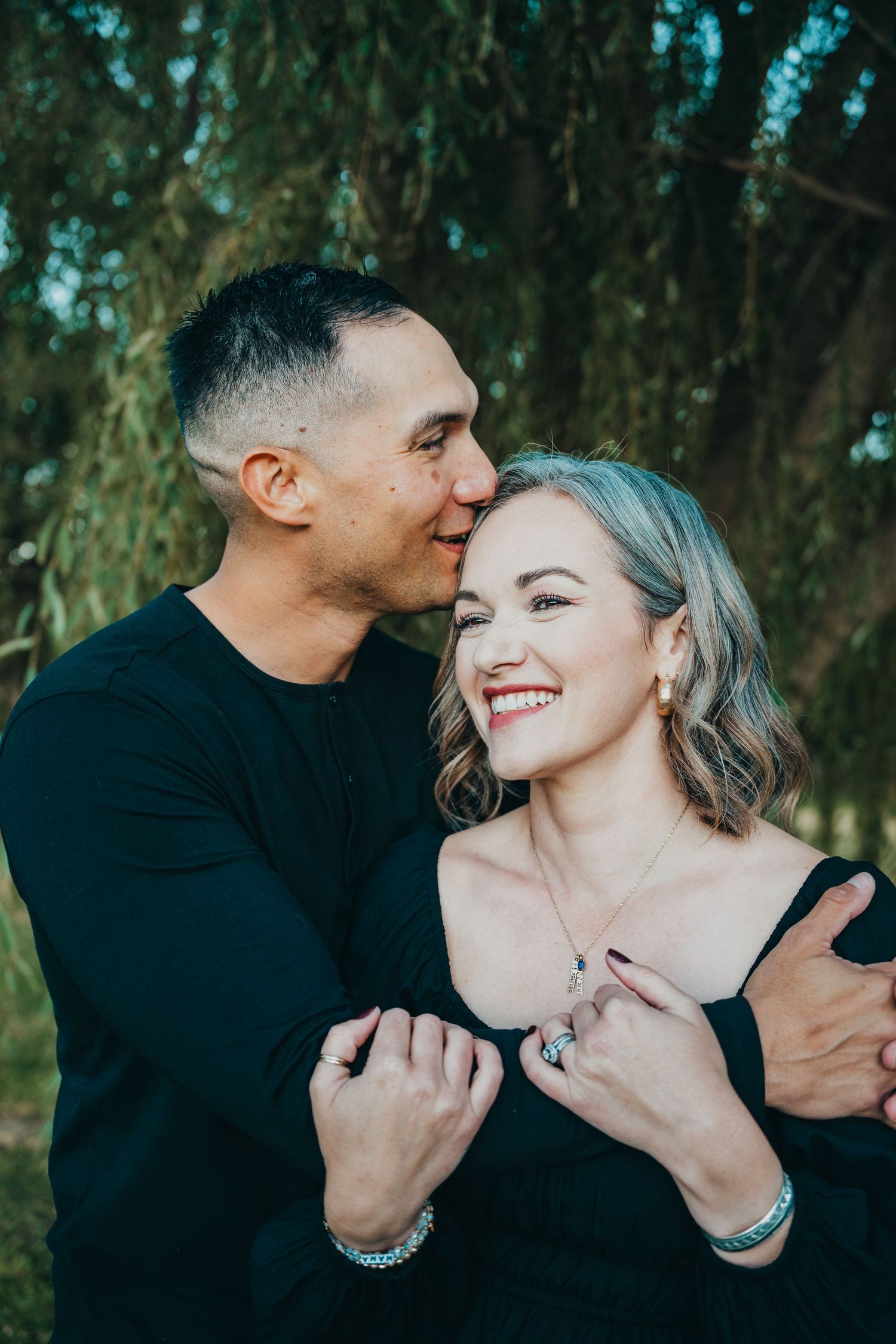 A loving couple embraces under a weeping willow tree, the man kissing the smiling woman on the cheek.