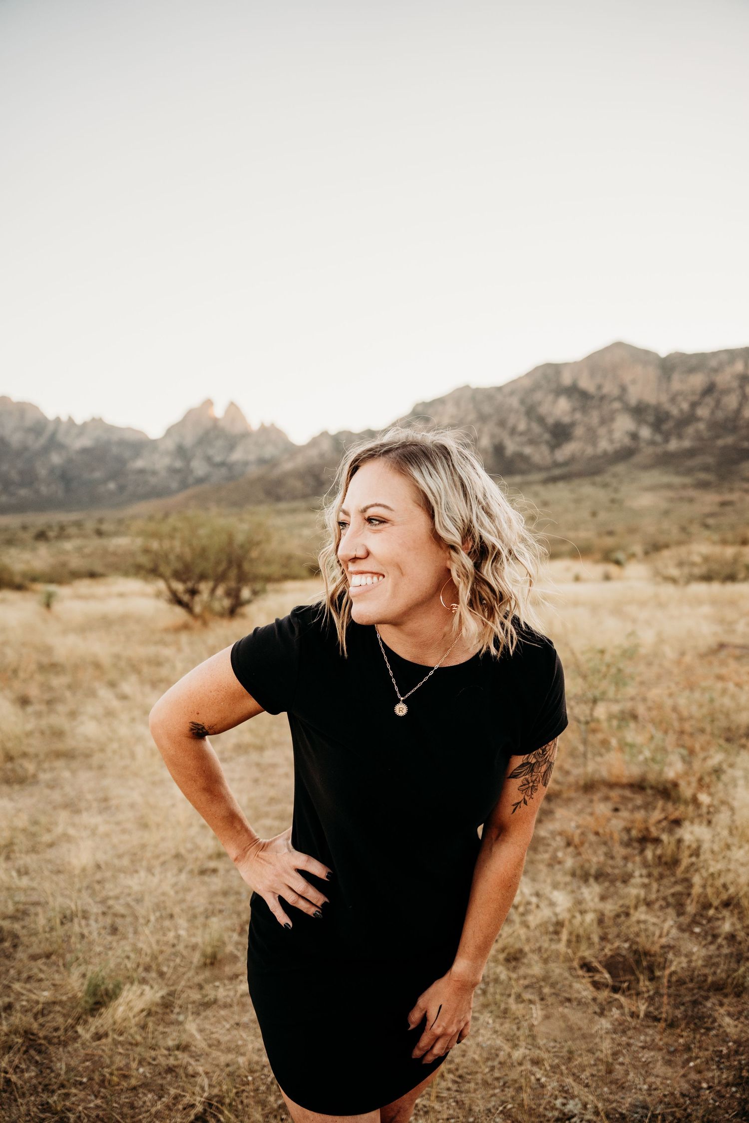 Smiling blonde woman in black dress poses in desert landscape with mountains.