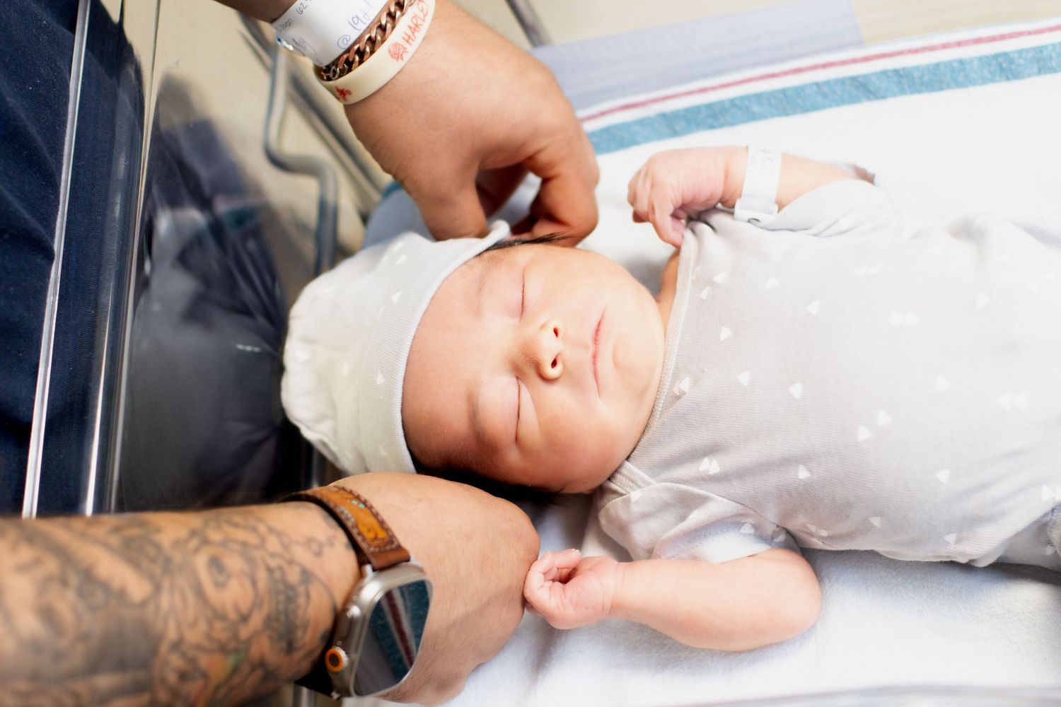 Tattooed father gently adjusting hat on sleeping newborn baby in hospital.