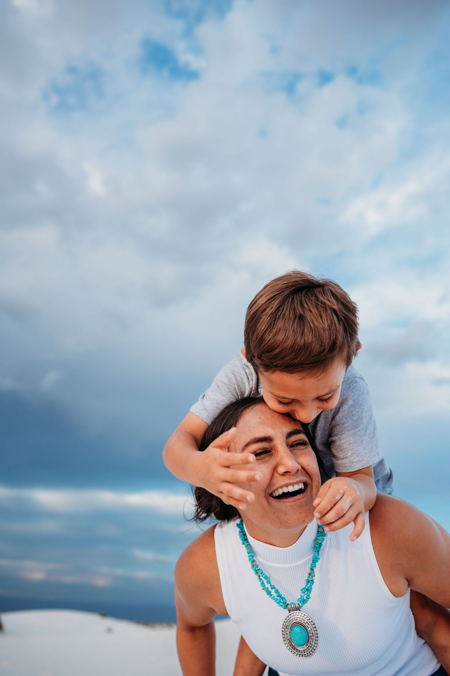 Laughing mother with turquoise necklace as young boy kisses her on white sandy beach.