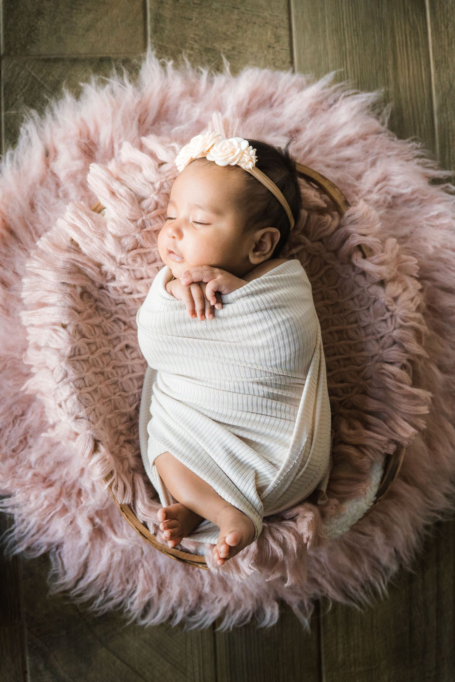 Overhead view of swaddled newborn baby girl with floral headband sleeping in pink fluffy basket.