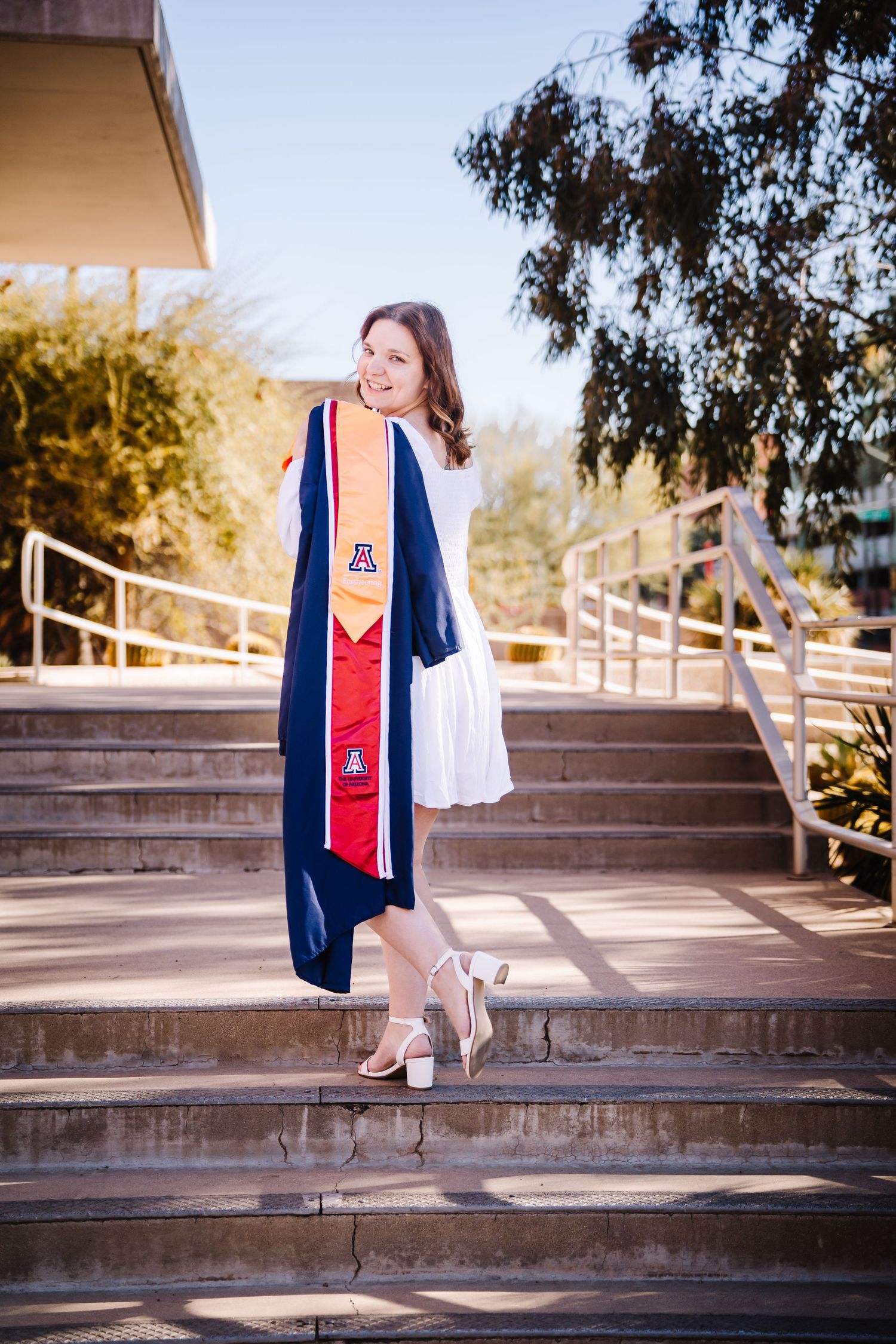University of Arizona graduate poses on campus steps in white dress, holding navy and red graduation regalia with UA logo.