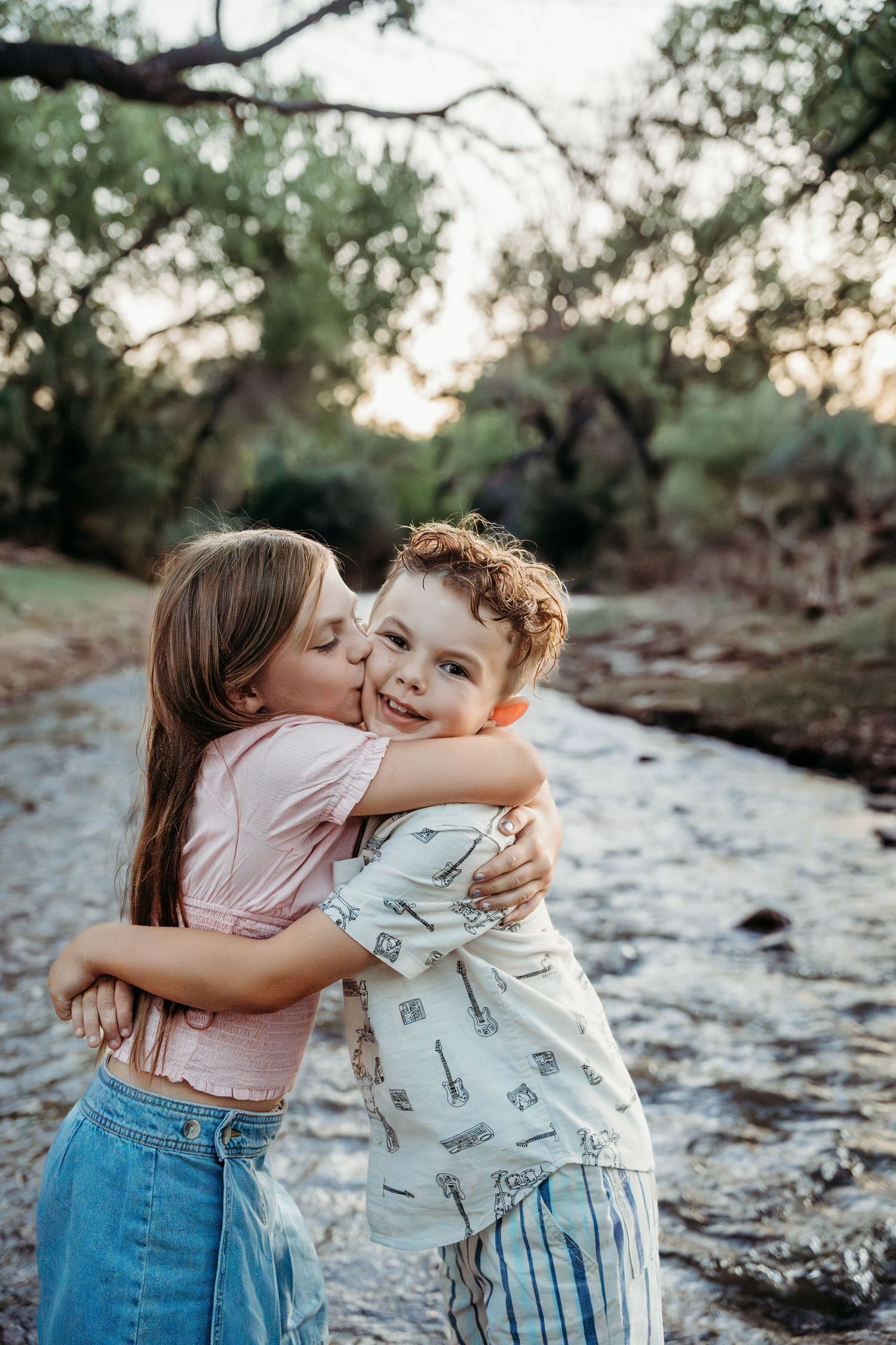A girl kisses her younger brother on the cheek while hugging him near a scenic creek.