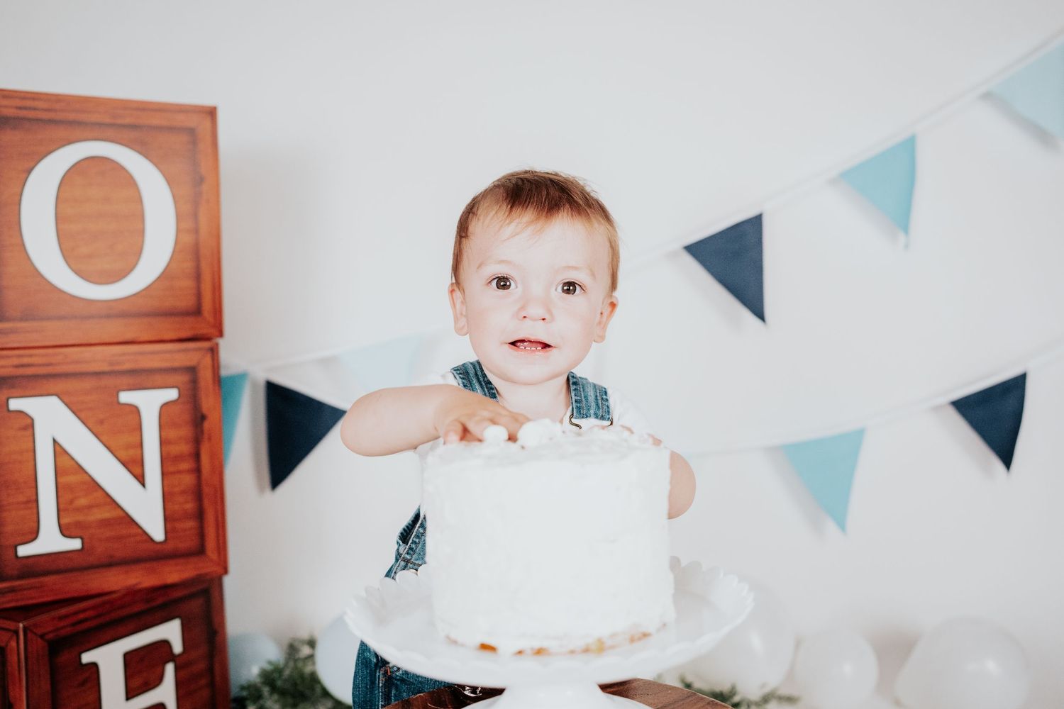 One-year-old boy in denim romper smiling behind white birthday cake with blue pennant flags.