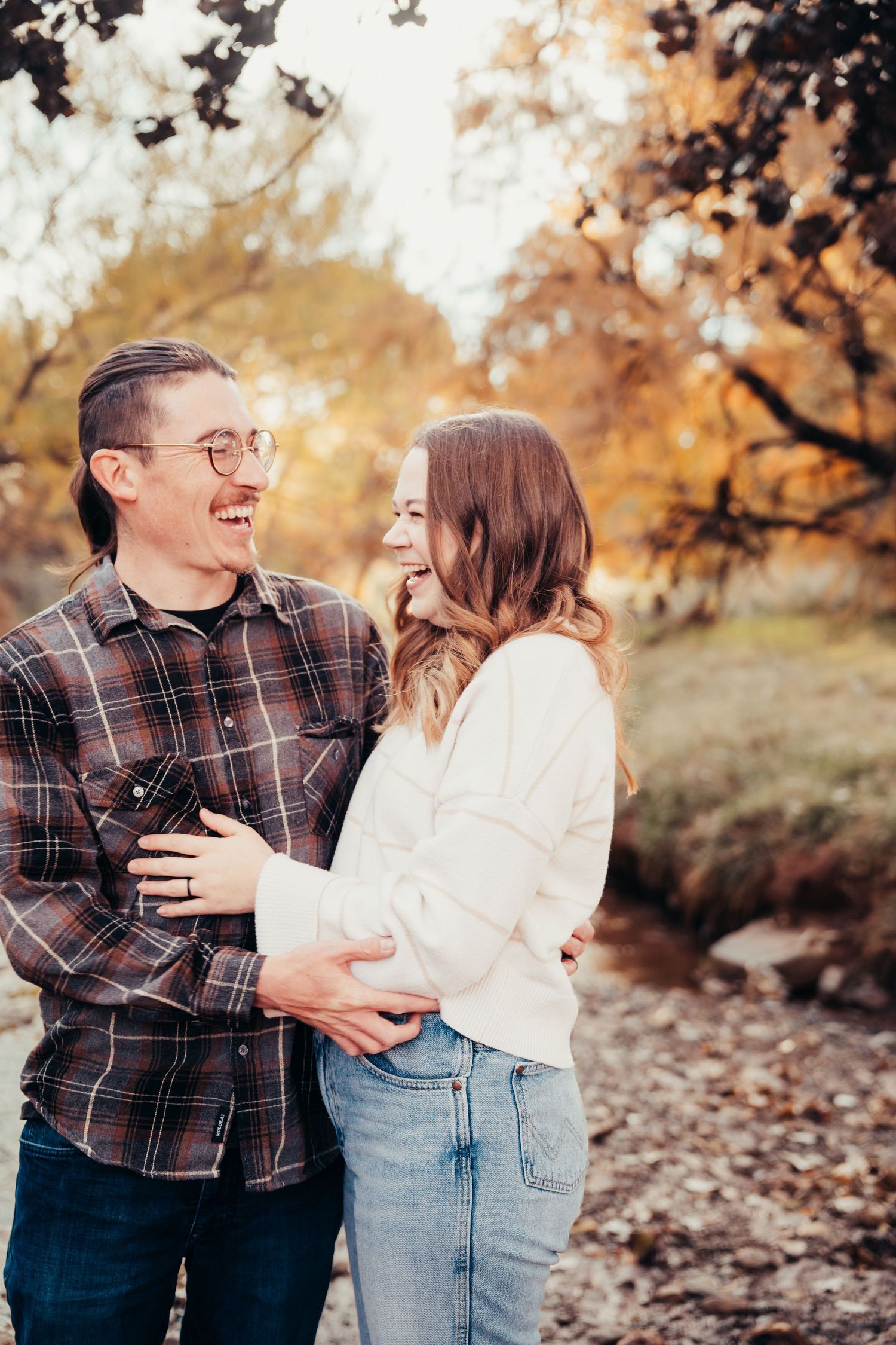 Laughing couple holding each other near a stream with golden fall trees.