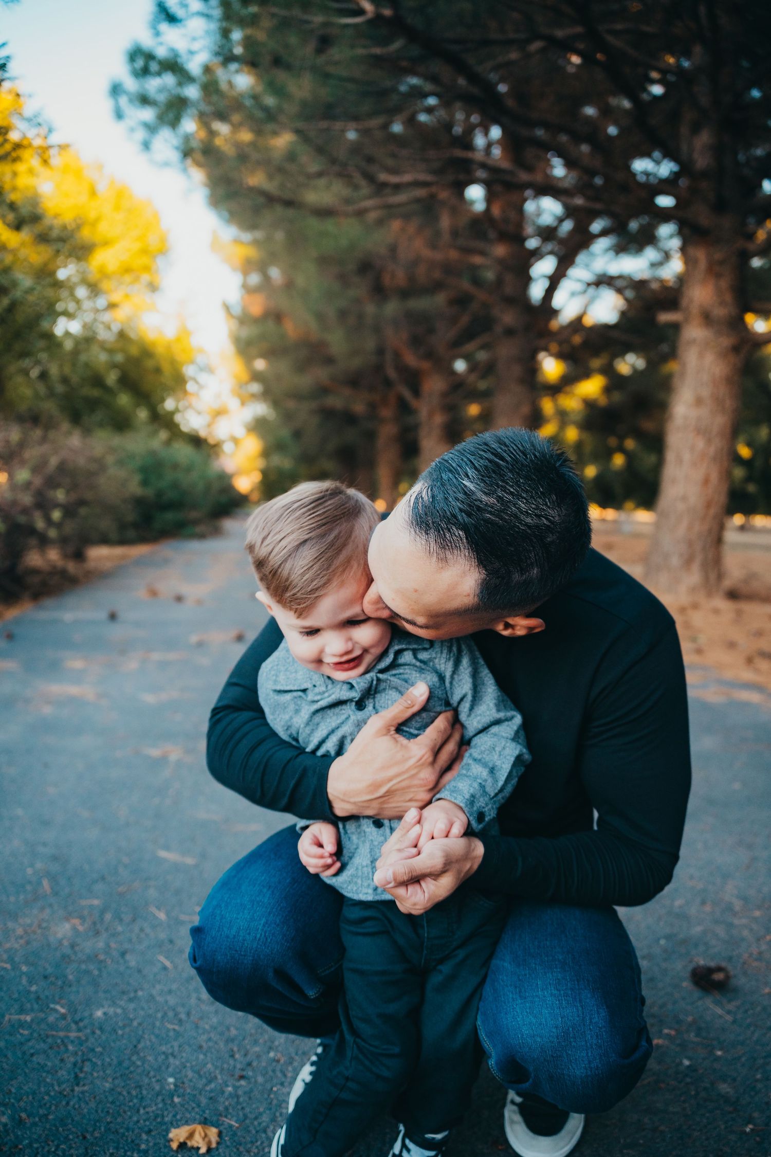 A loving father crouching down to kiss his smiling toddler son on a tree-lined park pathway.