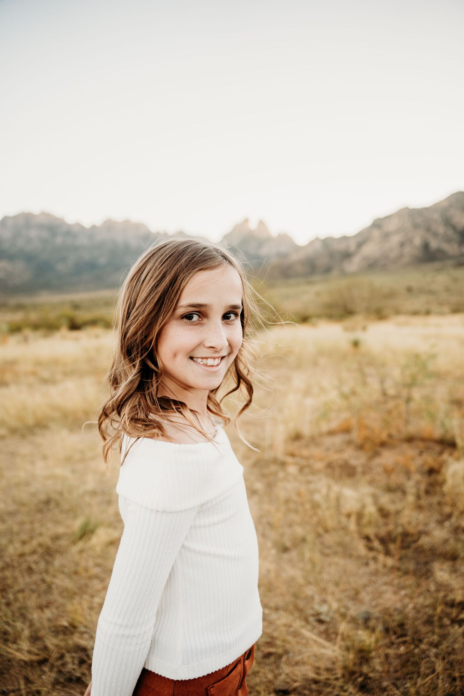 Smiling young girl in white off-shoulder top poses in golden field with mountains behind her.