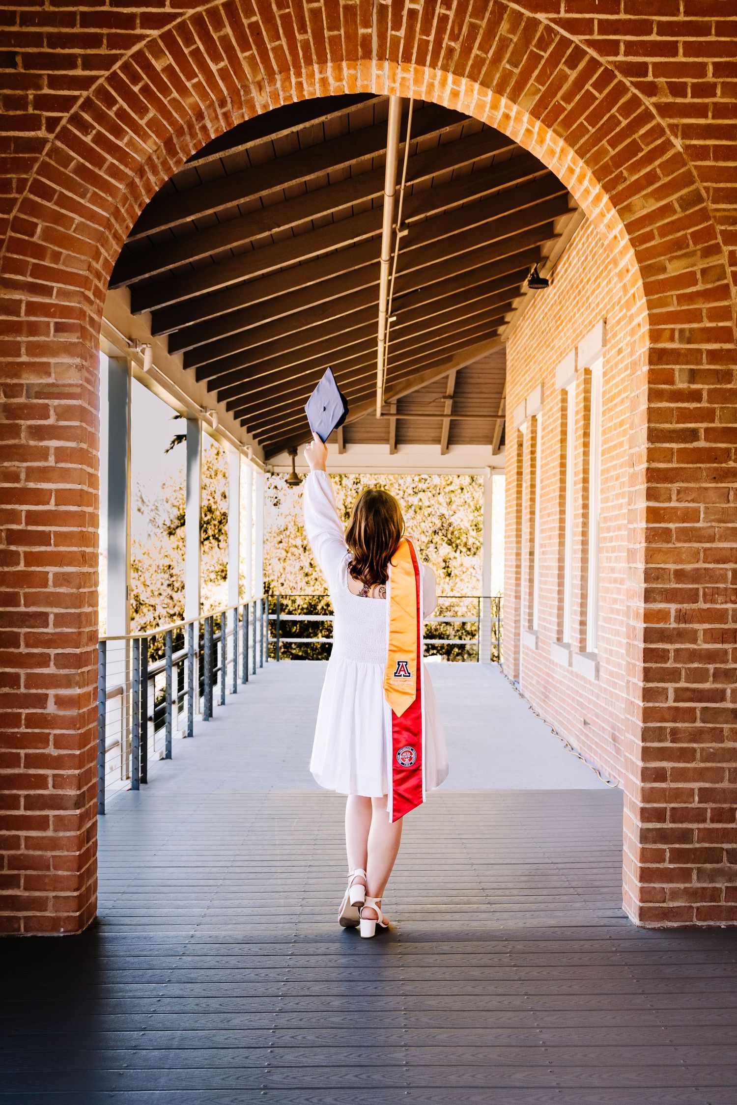 A graduate in white dress raises her cap under brick arches on a wooden walkway, celebrating her achievement.