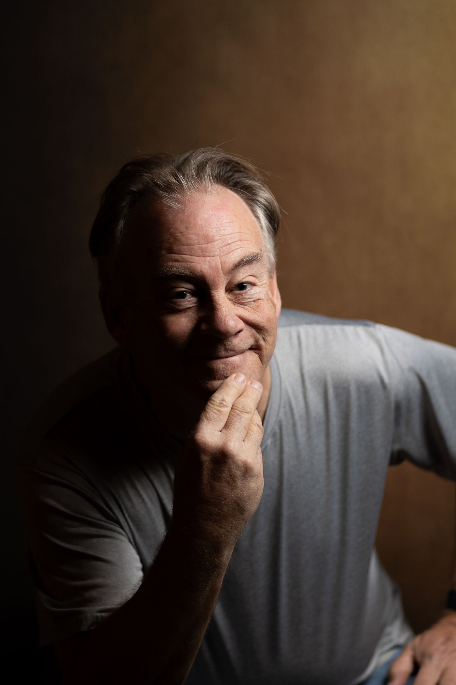 Older man in gray t-shirt smiling warmly with hand near chin against dramatic dark brown studio background.
