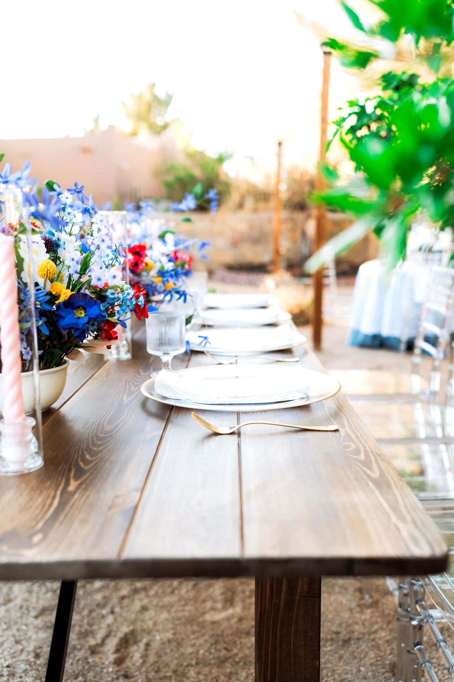 Outdoor wedding reception table with vibrant blue and red wildflower centerpieces, white plates, and gold utensils on wood.