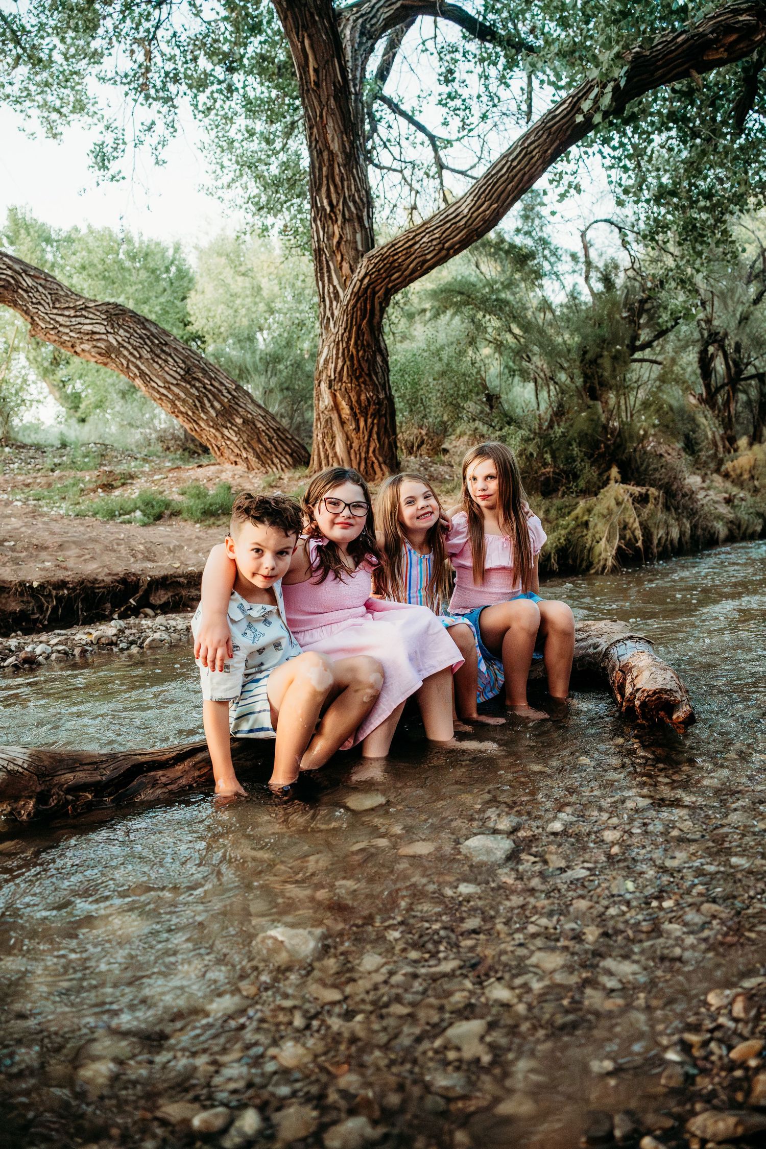 Four children sit on a log in a creek, smiling and splashing their feet in the water.