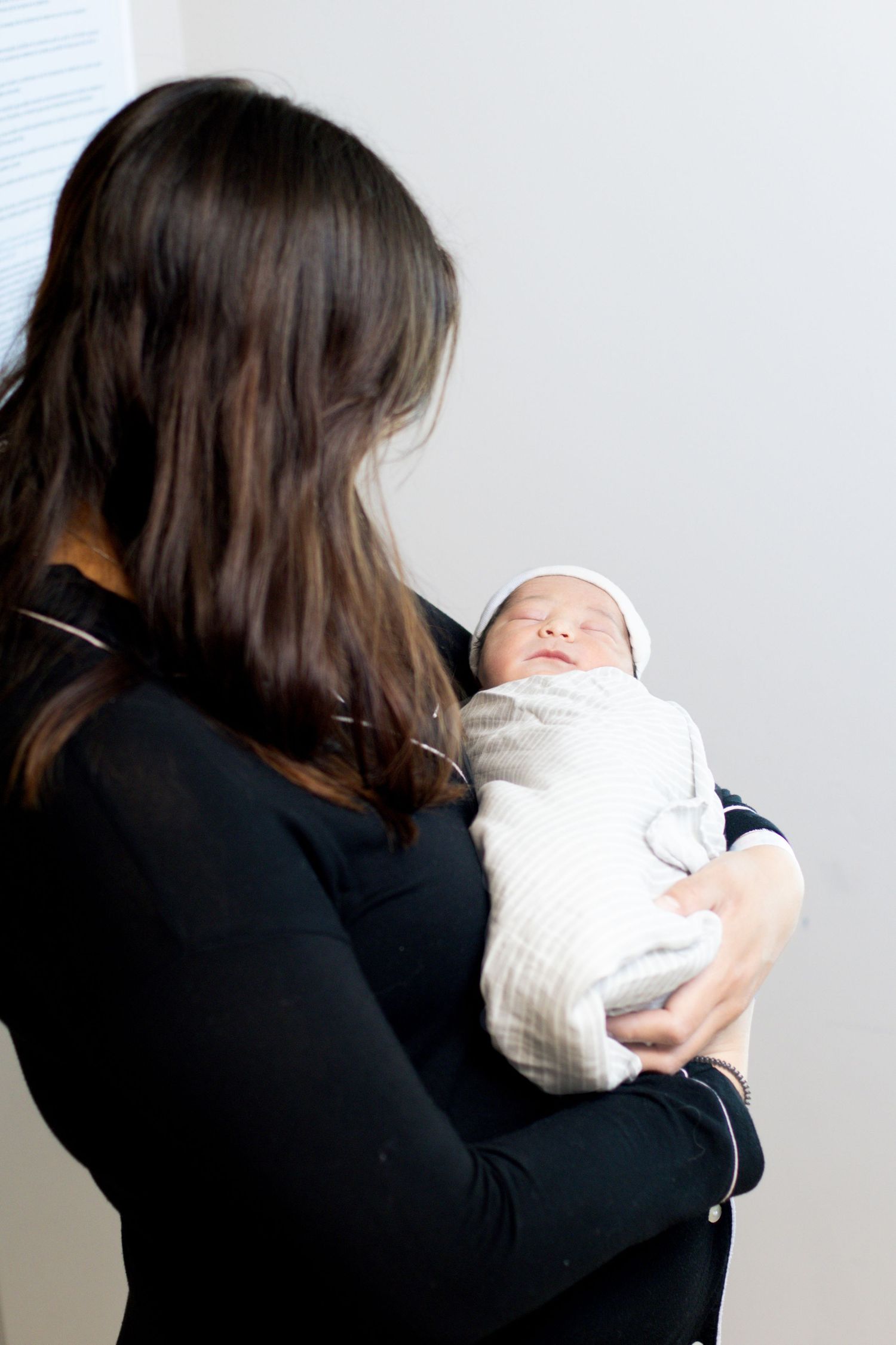 Mother in black outfit holding swaddled newborn baby from behind in bright hospital room.