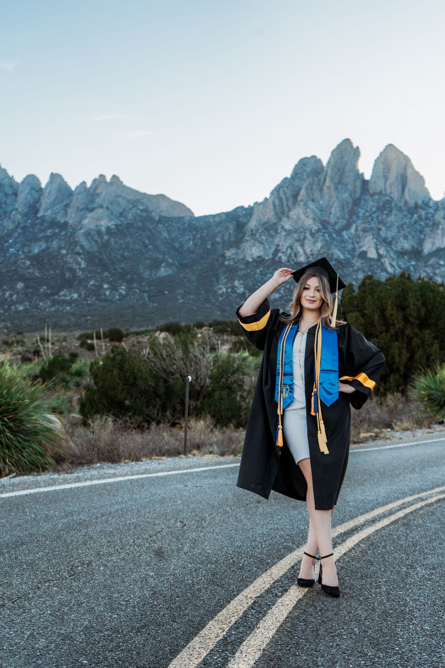 Graduate in black cap and gown with blue and gold stoles posing on road with rocky mountains behind.
