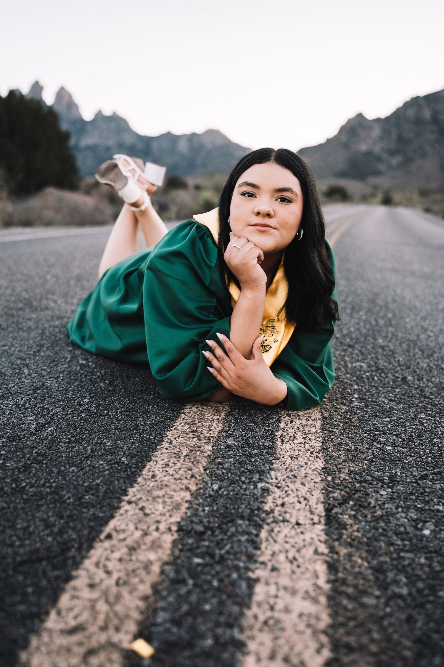 Graduate in green cap and gown lying on road centerline with scenic mountain landscape behind her.