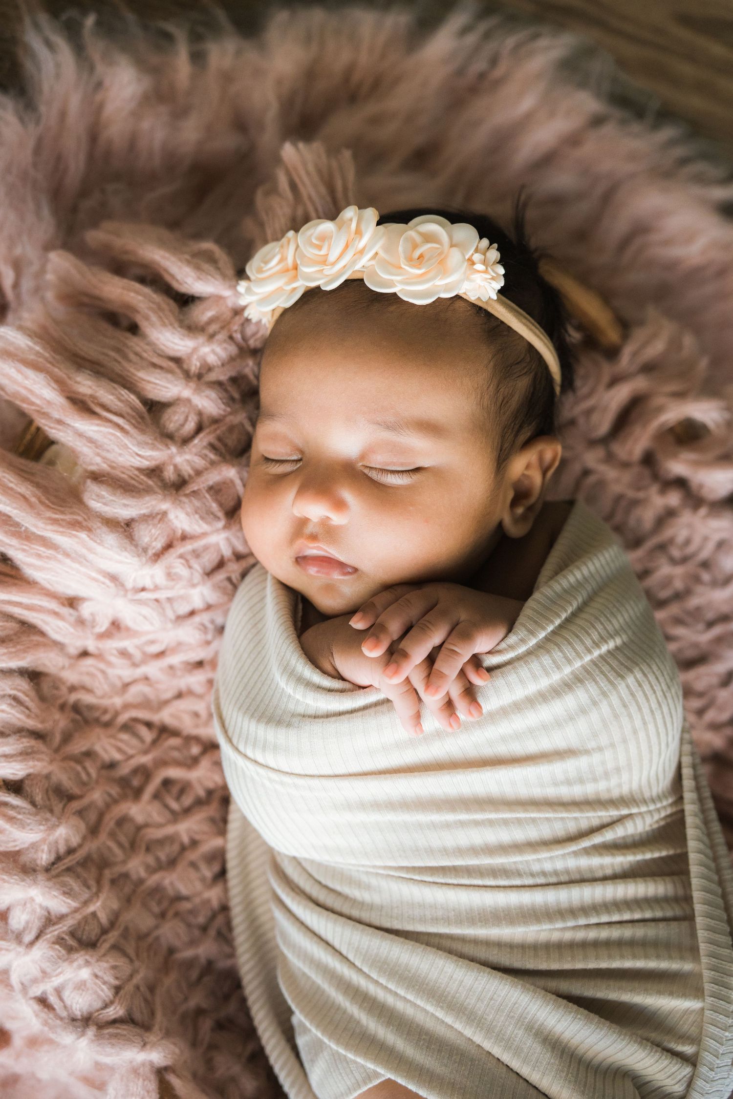 Sleeping newborn baby girl swaddled in cream wrap with floral headband on pink fluffy blanket.