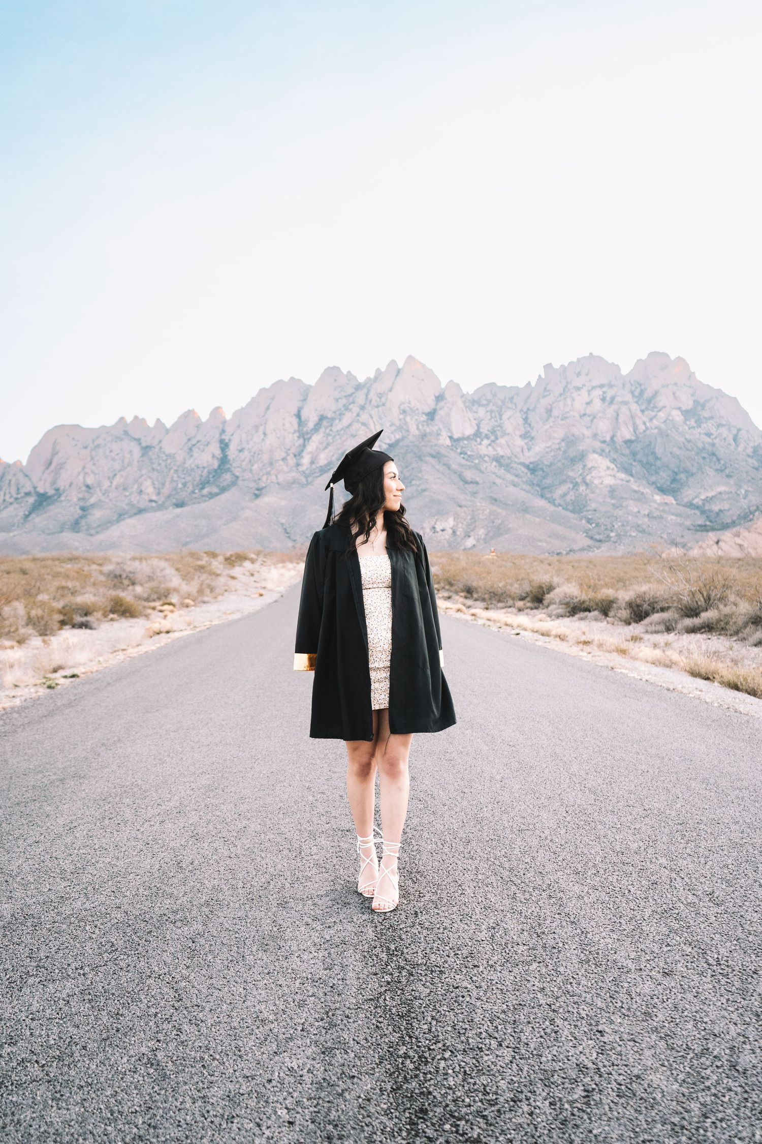 Female graduate in black cap and gown stands on an empty desert road with dramatic mountain backdrop.