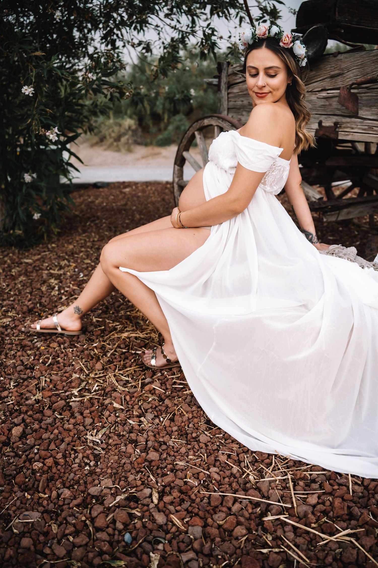 Pregnant woman in white dress and floral crown sits gracefully beside an antique wooden wagon.