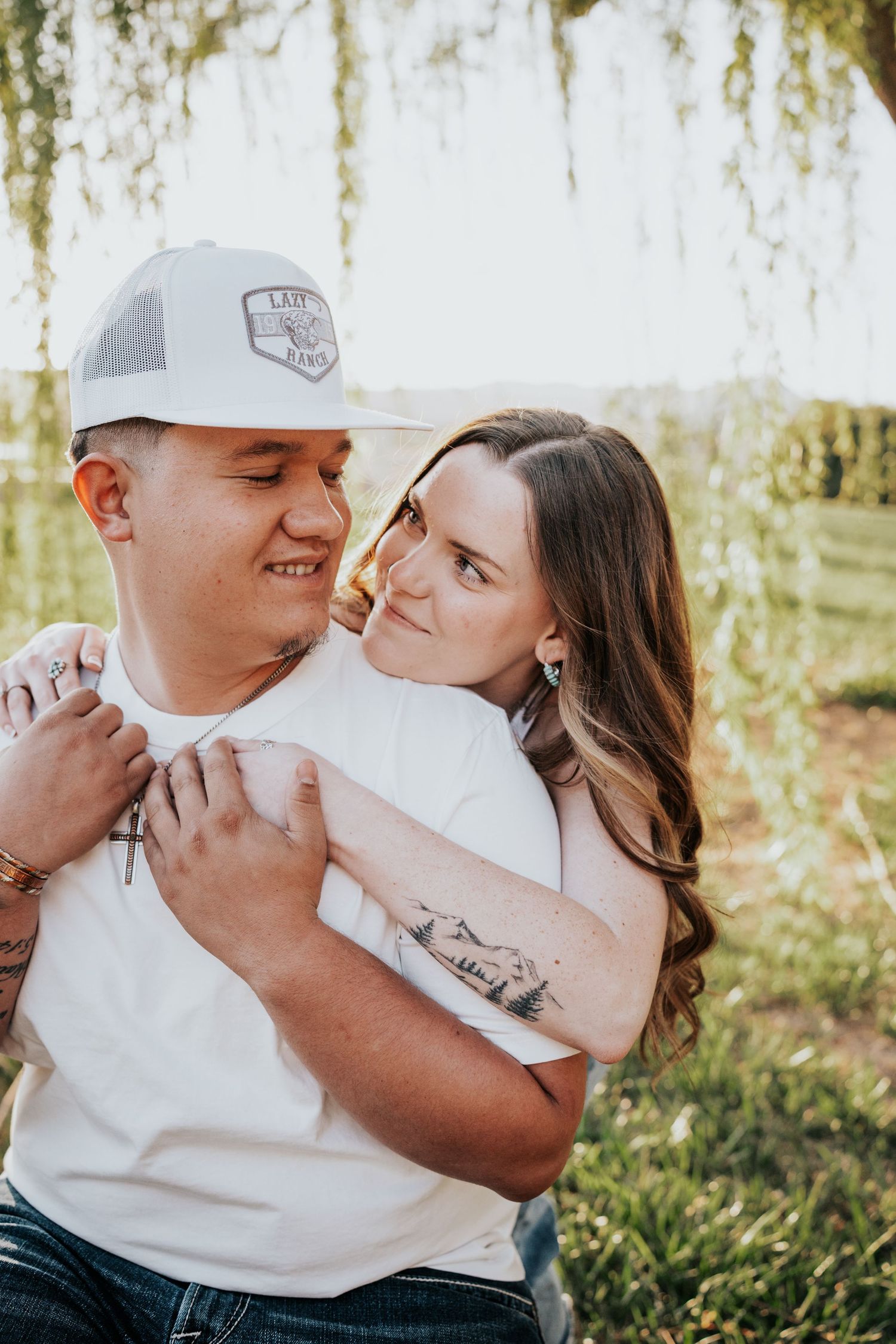 Woman with mountain tattoo hugging smiling man from behind in a lush green outdoor setting.