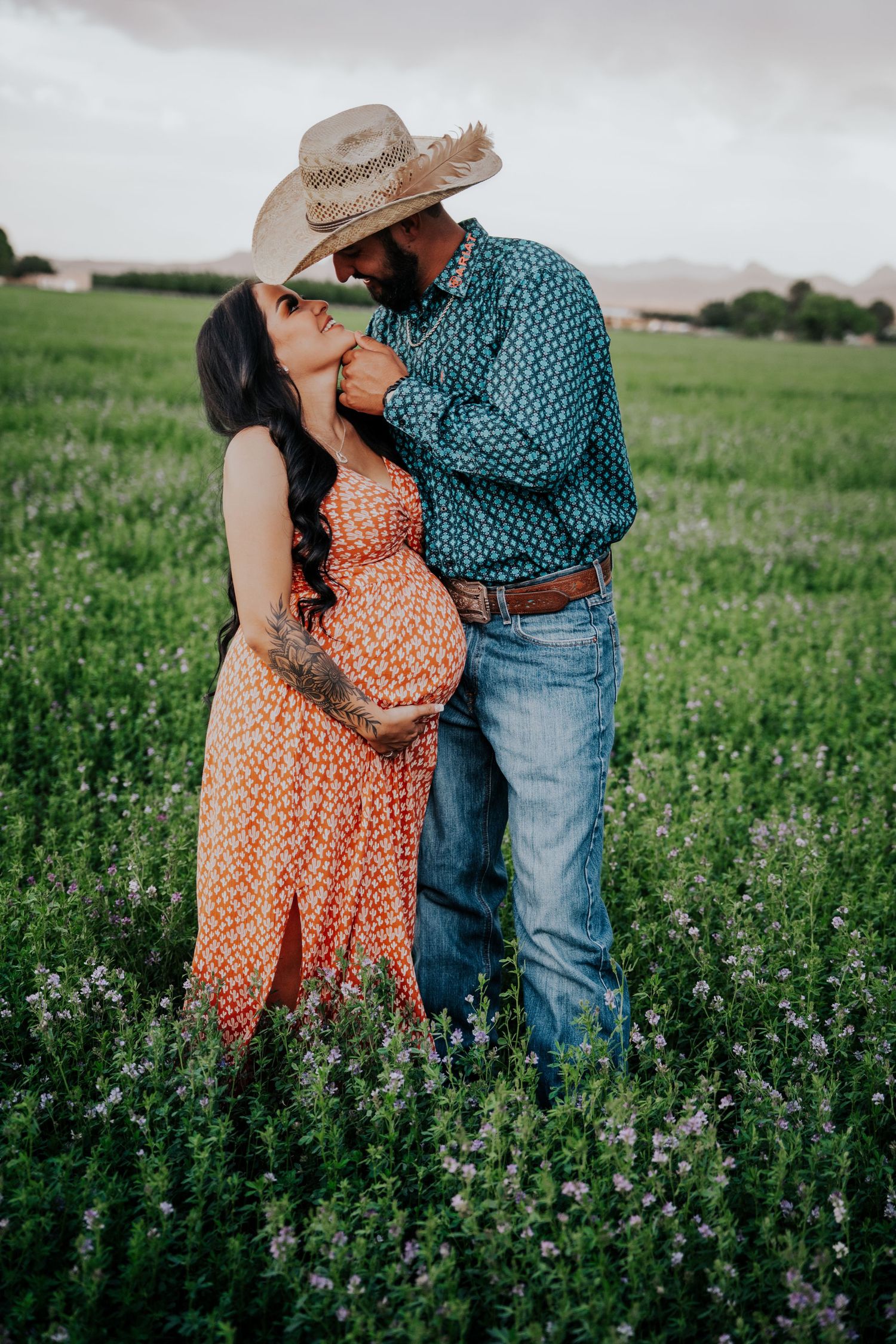 Pregnant couple kissing in blooming field, man in straw cowboy hat and teal shirt, woman in orange floral dress.
