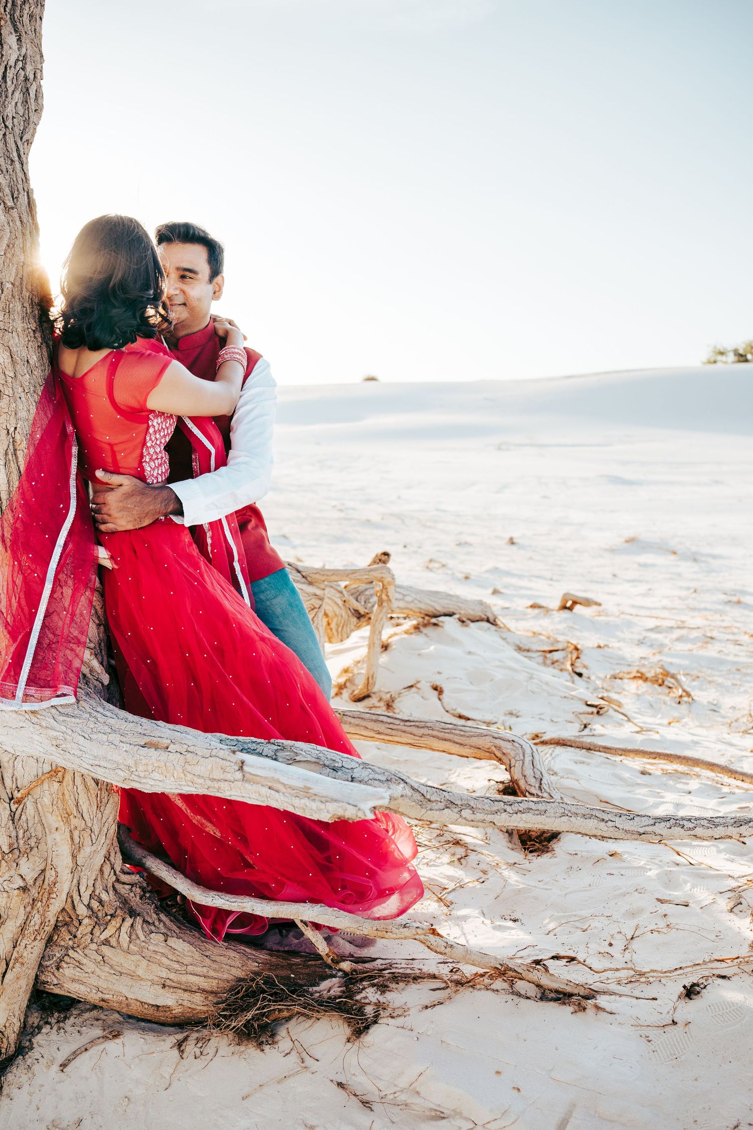 Couple in red Indian traditional wear embracing near a tree on bright white sandy desert dunes.