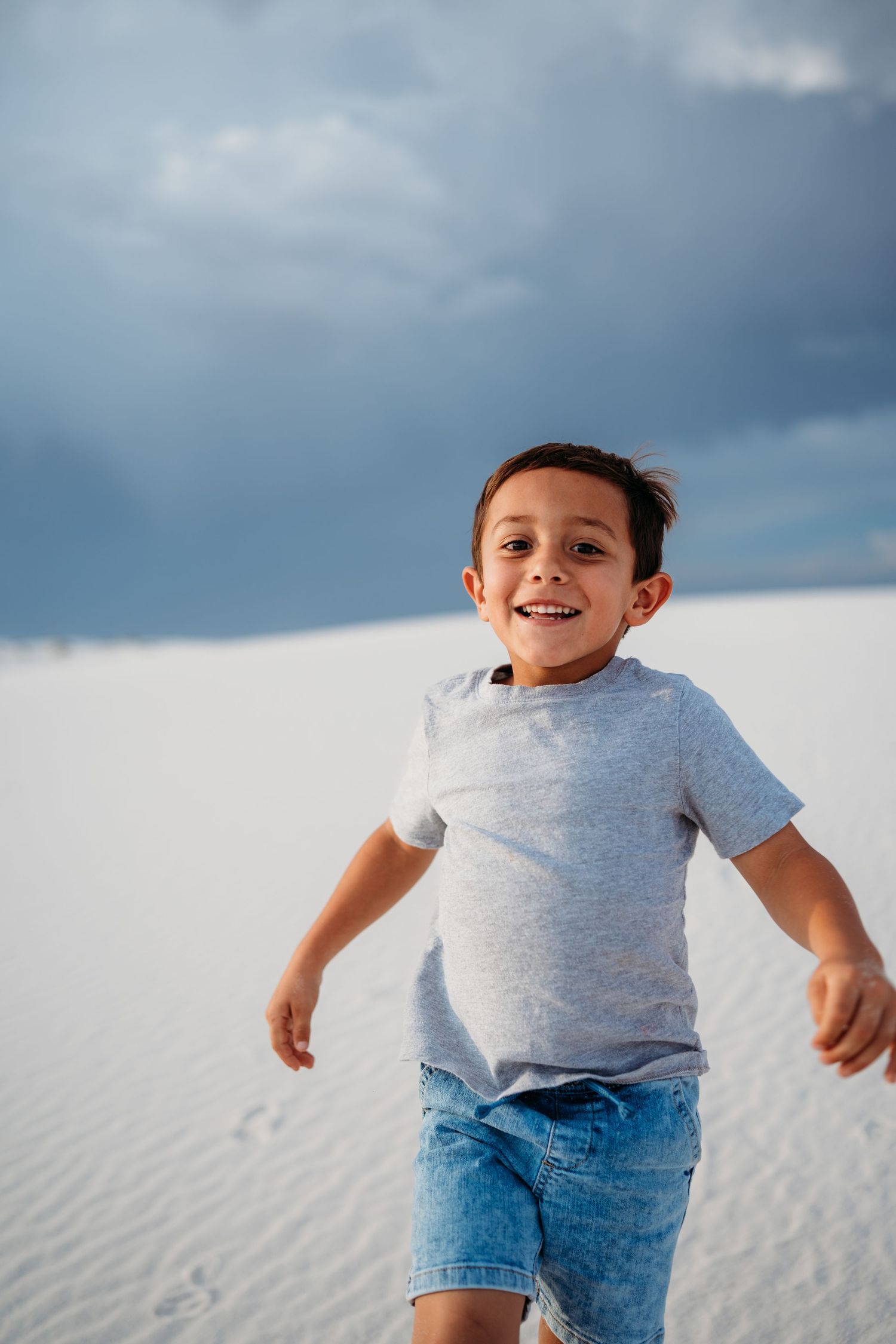 A happy young boy in a gray shirt and denim shorts runs joyfully across white sand dunes.