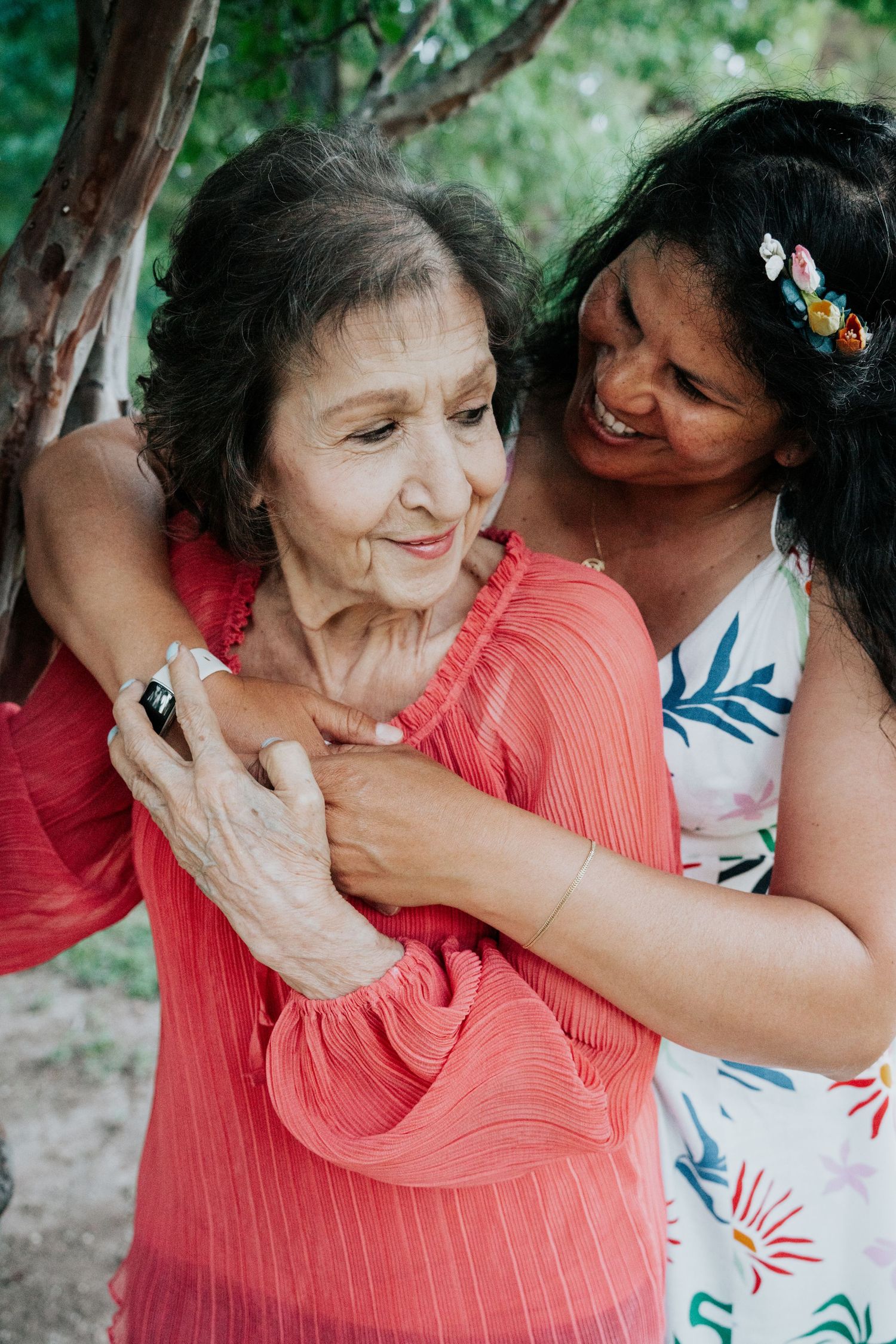 A smiling woman warmly embraces an elderly woman in a red blouse beneath a tree outdoors.