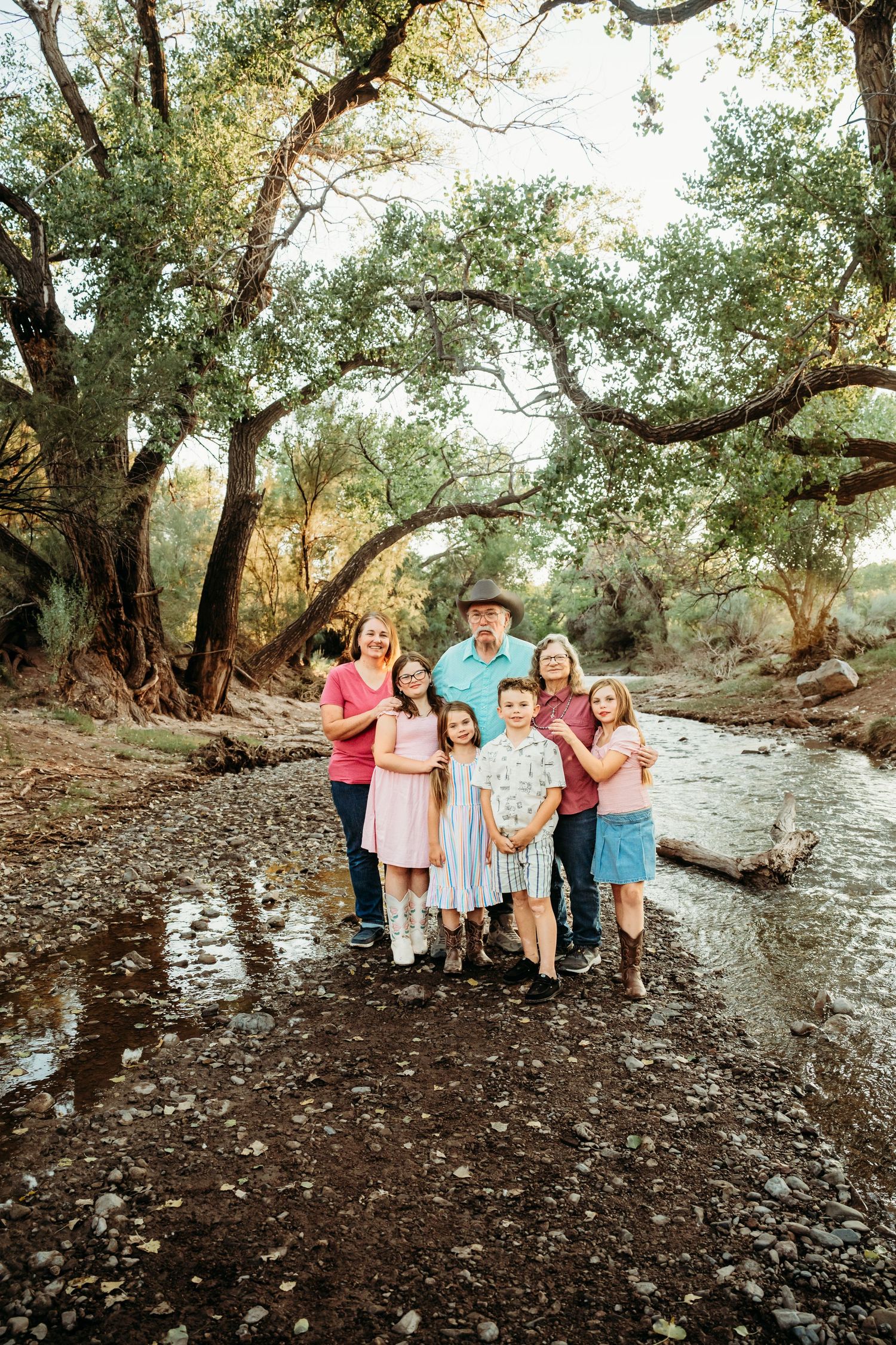 A large multigenerational family smiles together along a sunlit rocky creek surrounded by tall trees.