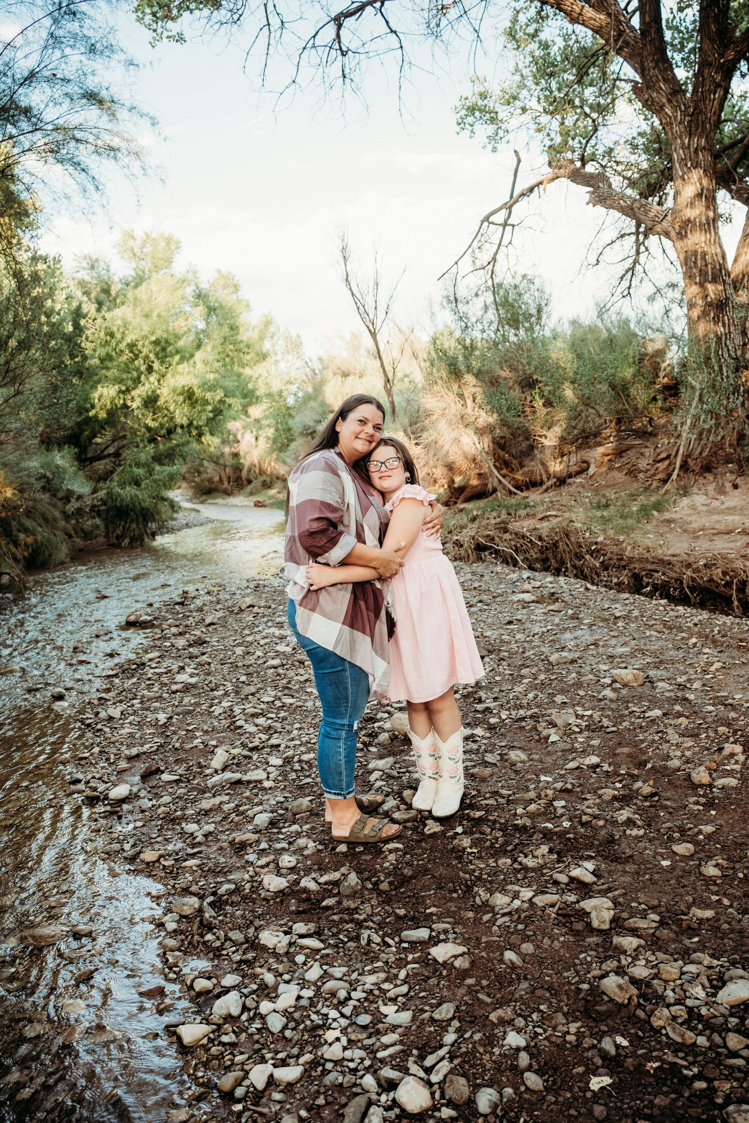 Mother and young daughter in pink dress hugging warmly by a scenic creek in autumn nature setting.
