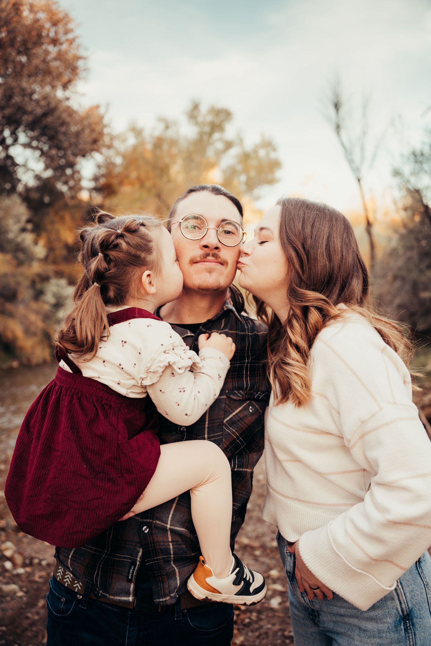 Sweet family moment with young parents both kissing toddler daughter on cheeks in autumn outdoor setting.