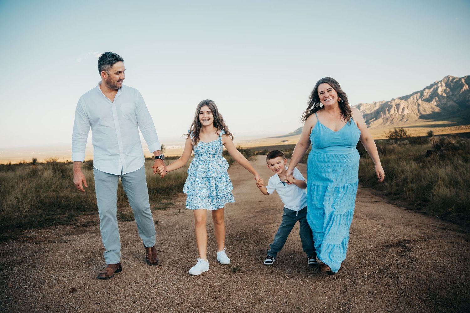 A happy family of four holds hands and walks along a dirt path during golden hour.
