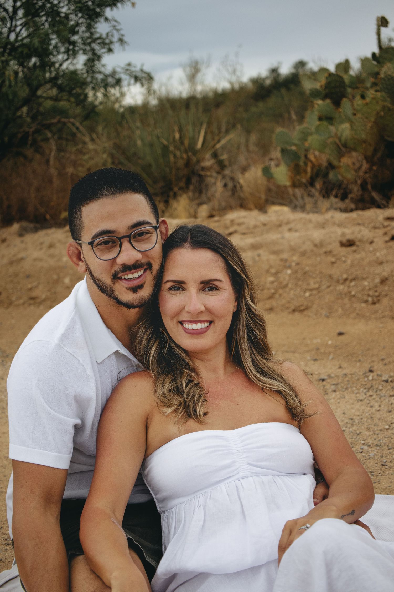Happy couple posing together in white outfits seated in a desert setting with cacti.