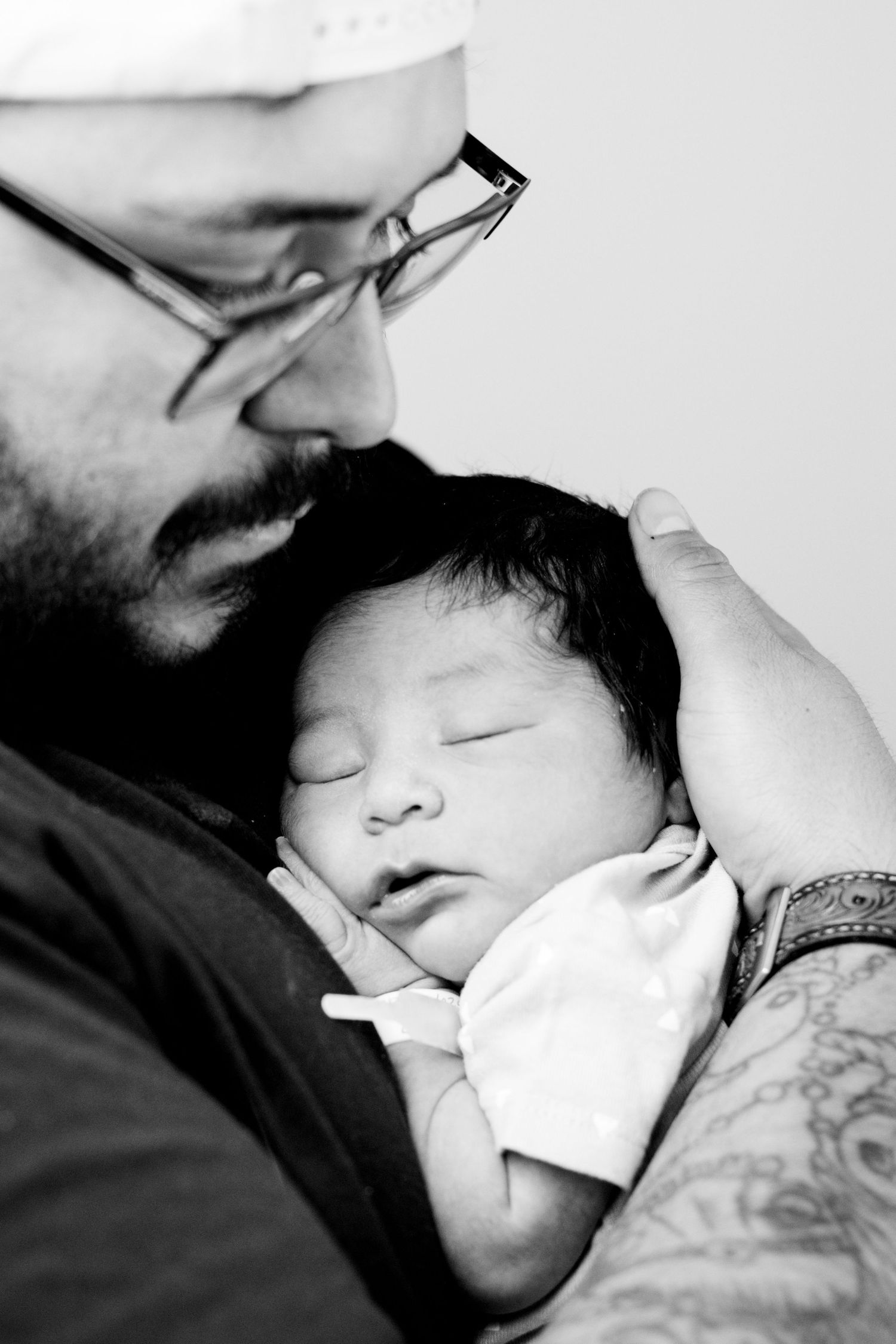 Black and white photo of tattooed father with glasses cradling sleeping newborn.