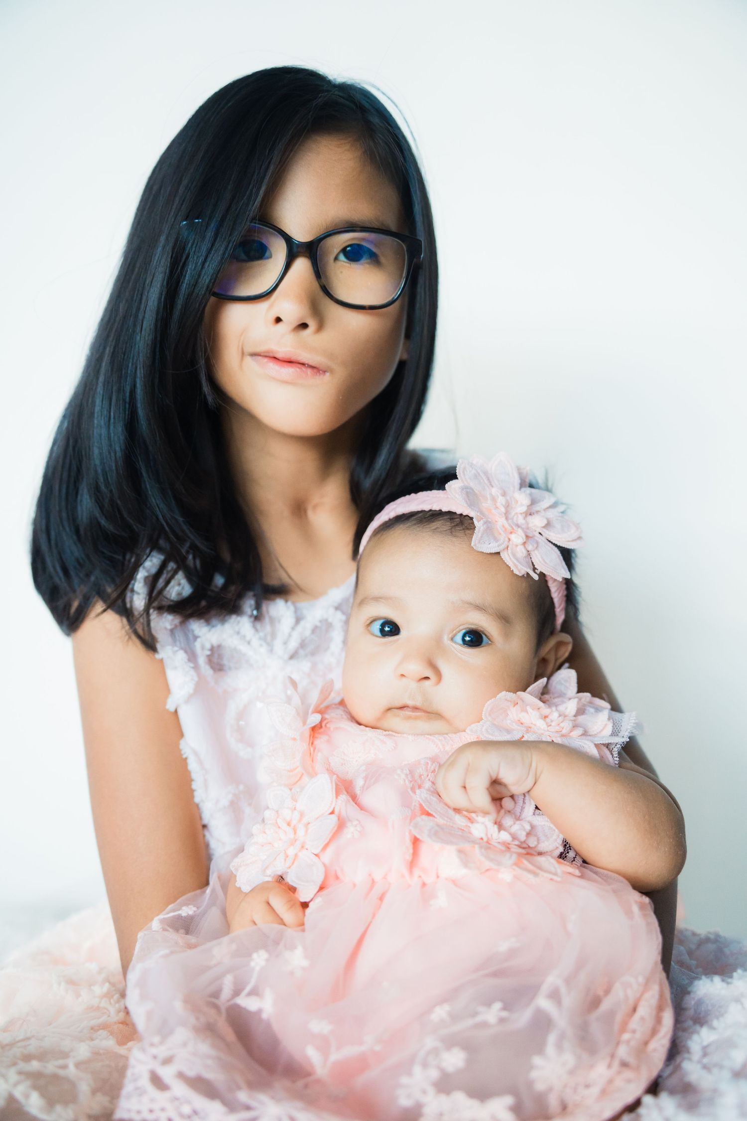 Young girl with glasses holding newborn baby sister in matching pink lace dresses portrait.