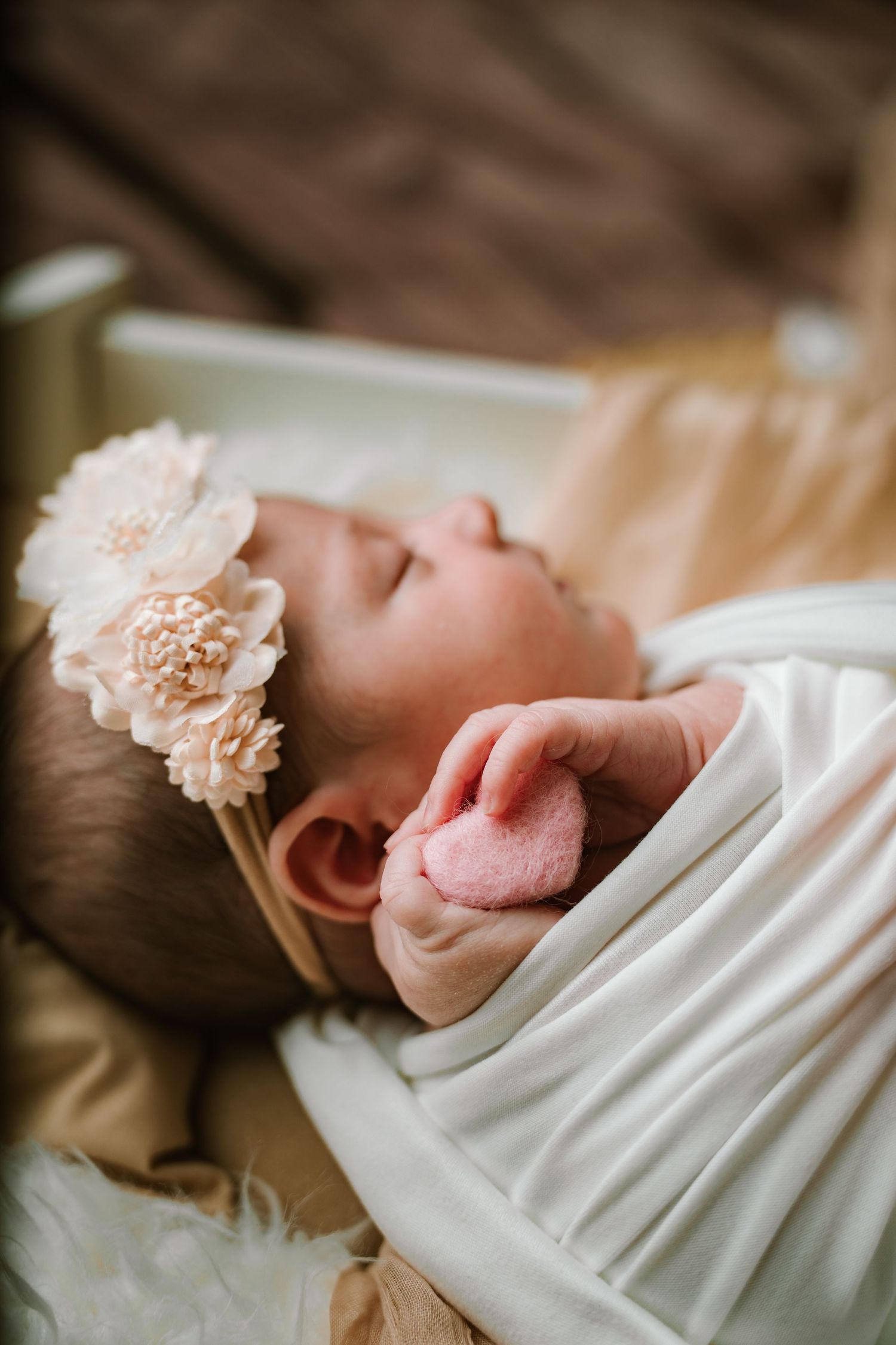 Newborn baby girl sleeping in white swaddle holding pink heart, wearing floral headband in soft light.