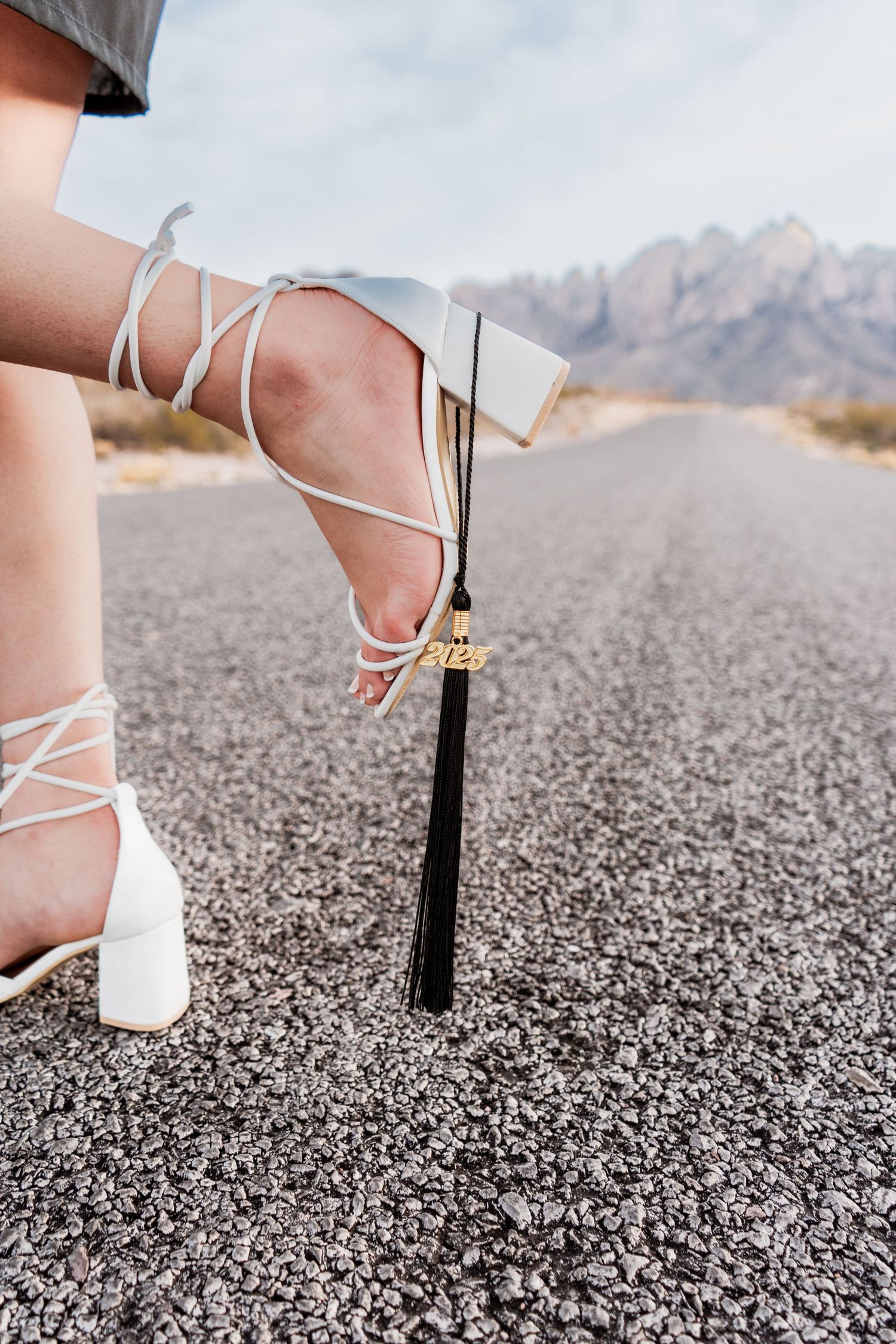 Close-up of white lace-up heels with graduation tassel on asphalt road with mountains behind.