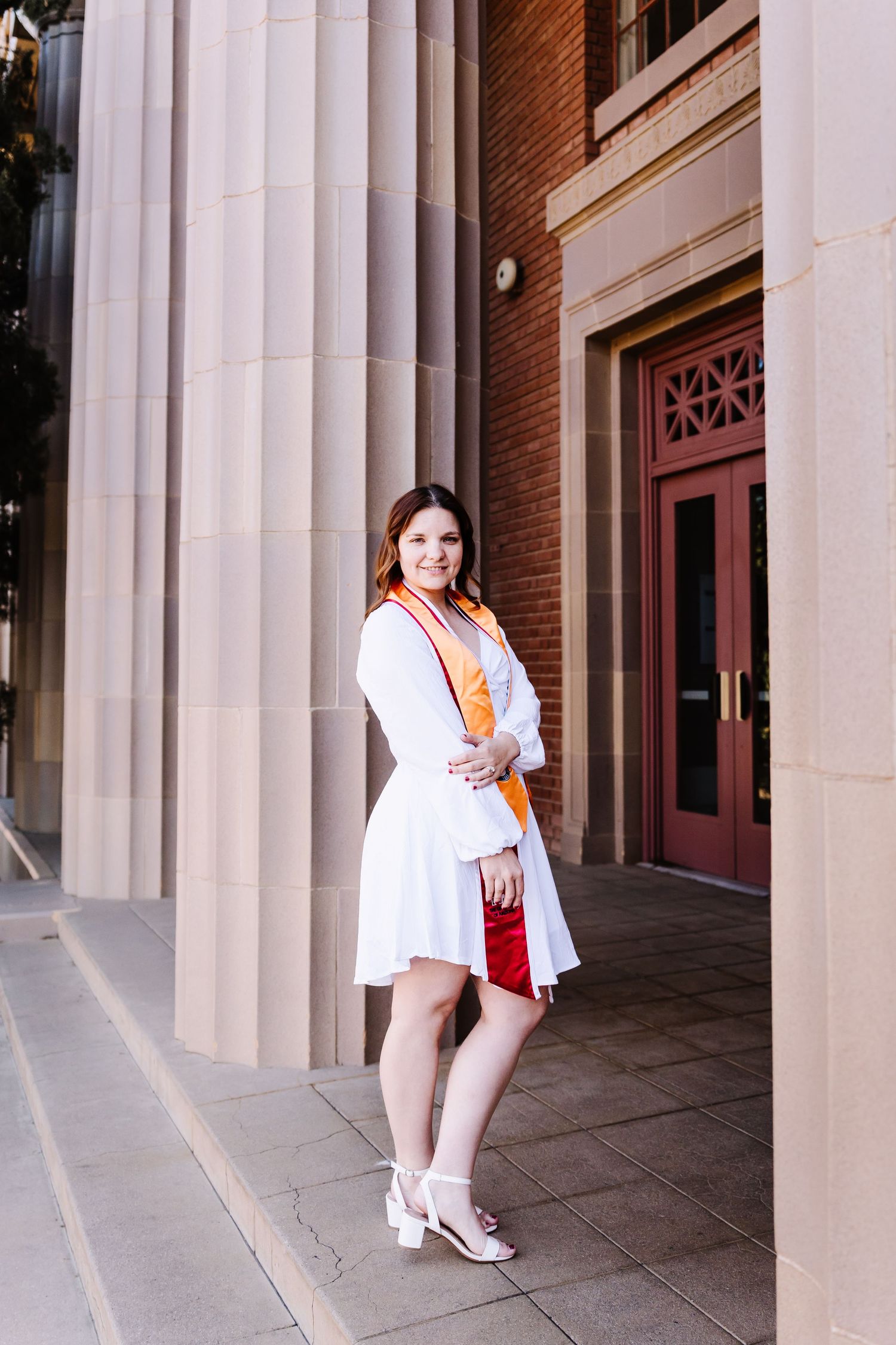 Graduate in white dress and orange sash poses between columns at university entrance.