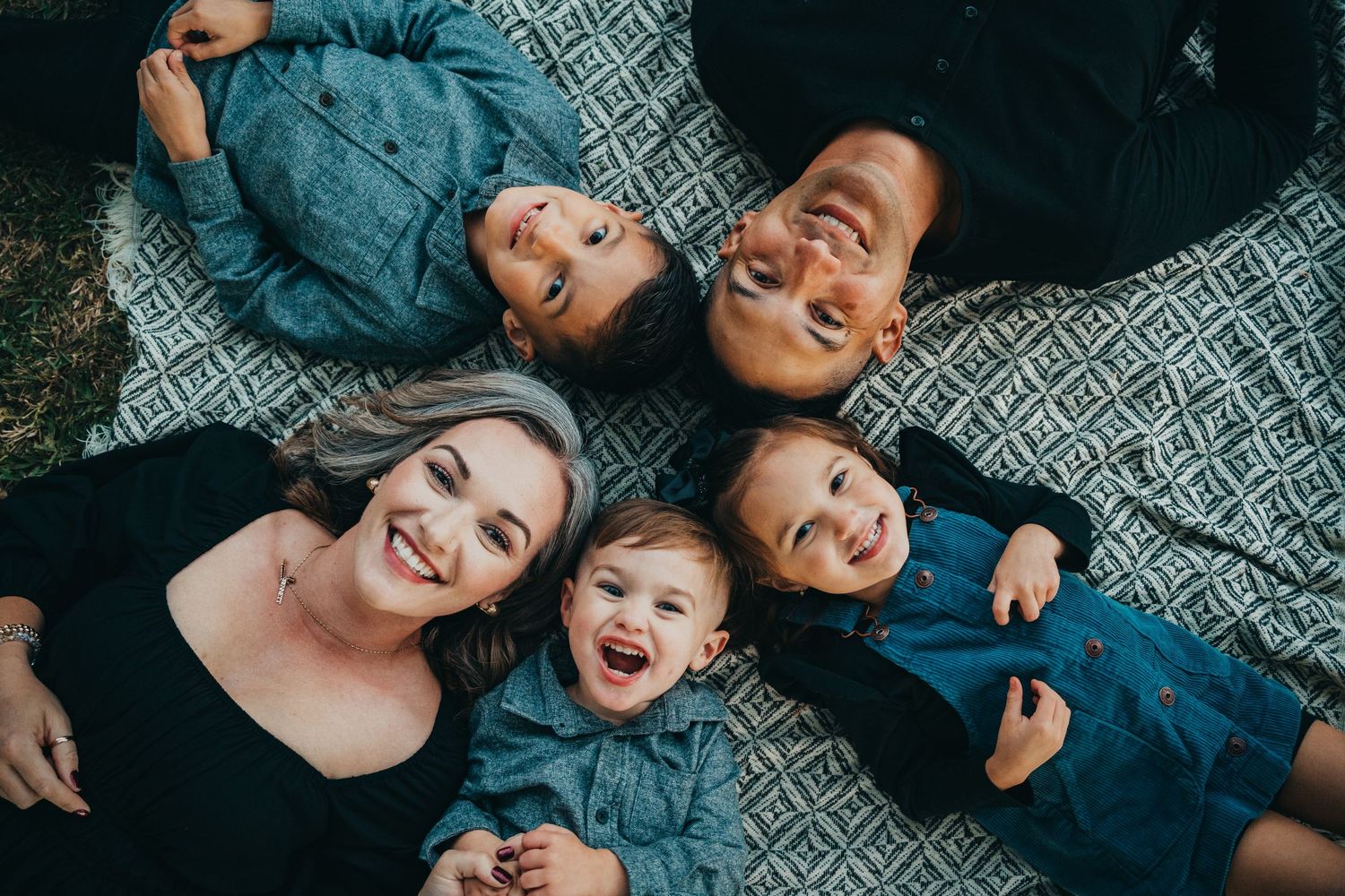 Joyful family of five lying in a circle on a blanket, smiling up at the camera in coordinated dark outfits.